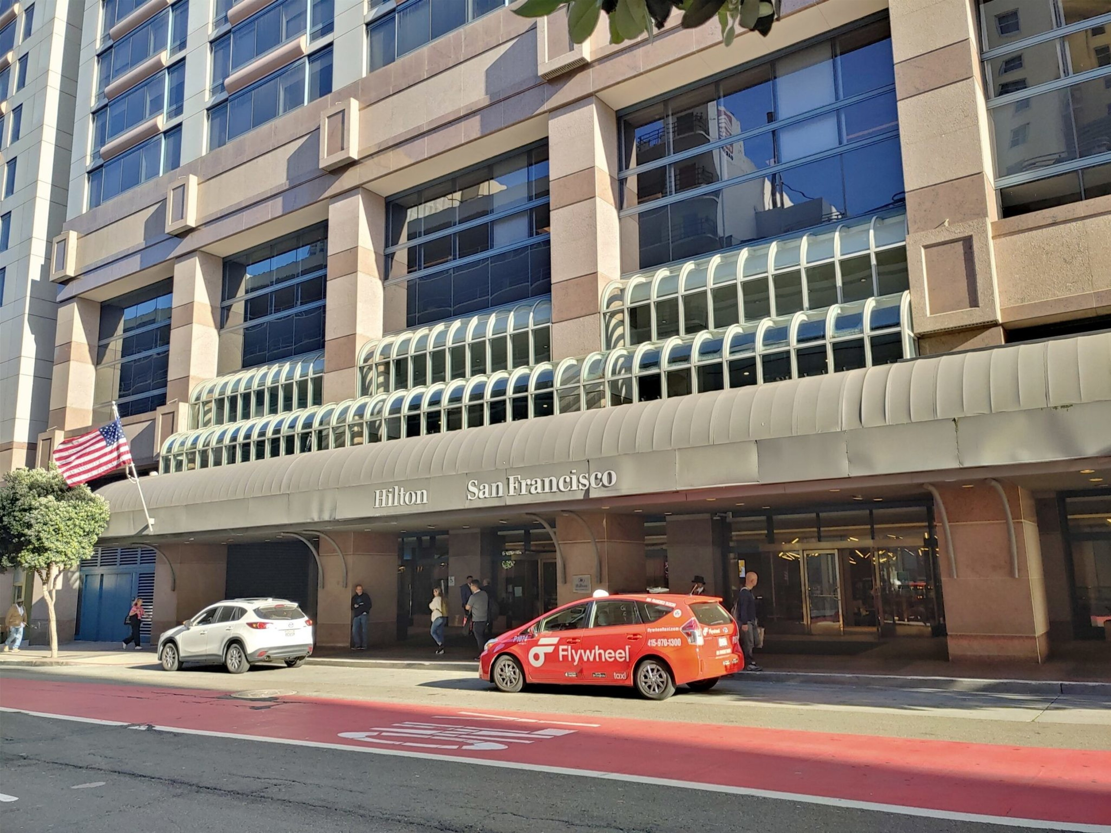 The image shows the entrance of the Hilton San Francisco hotel. A red taxi with "Flywheel" branding and several pedestrians are in front. An American flag is on the left.