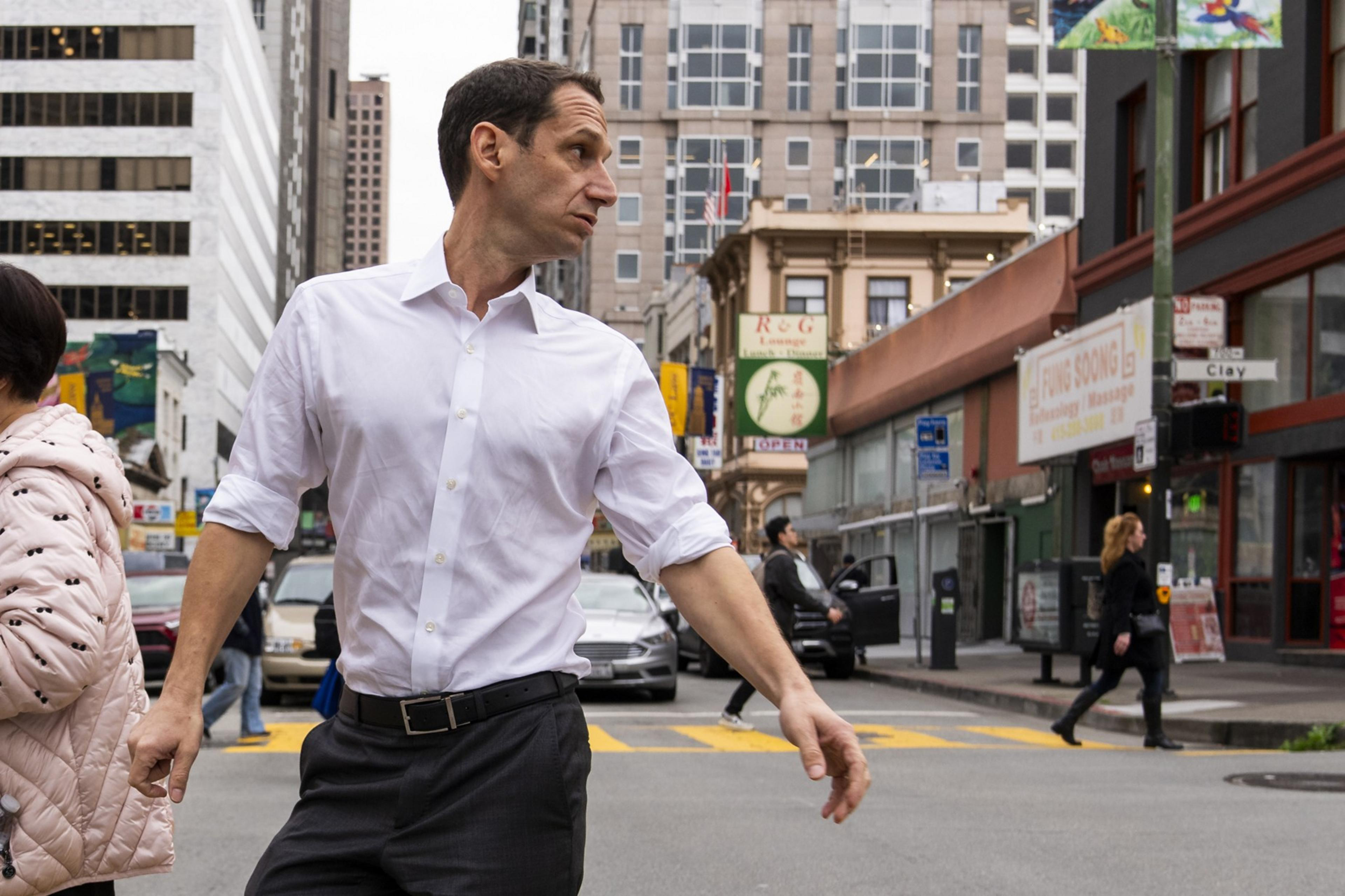 A man in a white shirt crosses a busy city street, turning his head to the side. Buildings, people walking, and cars are visible in the background.