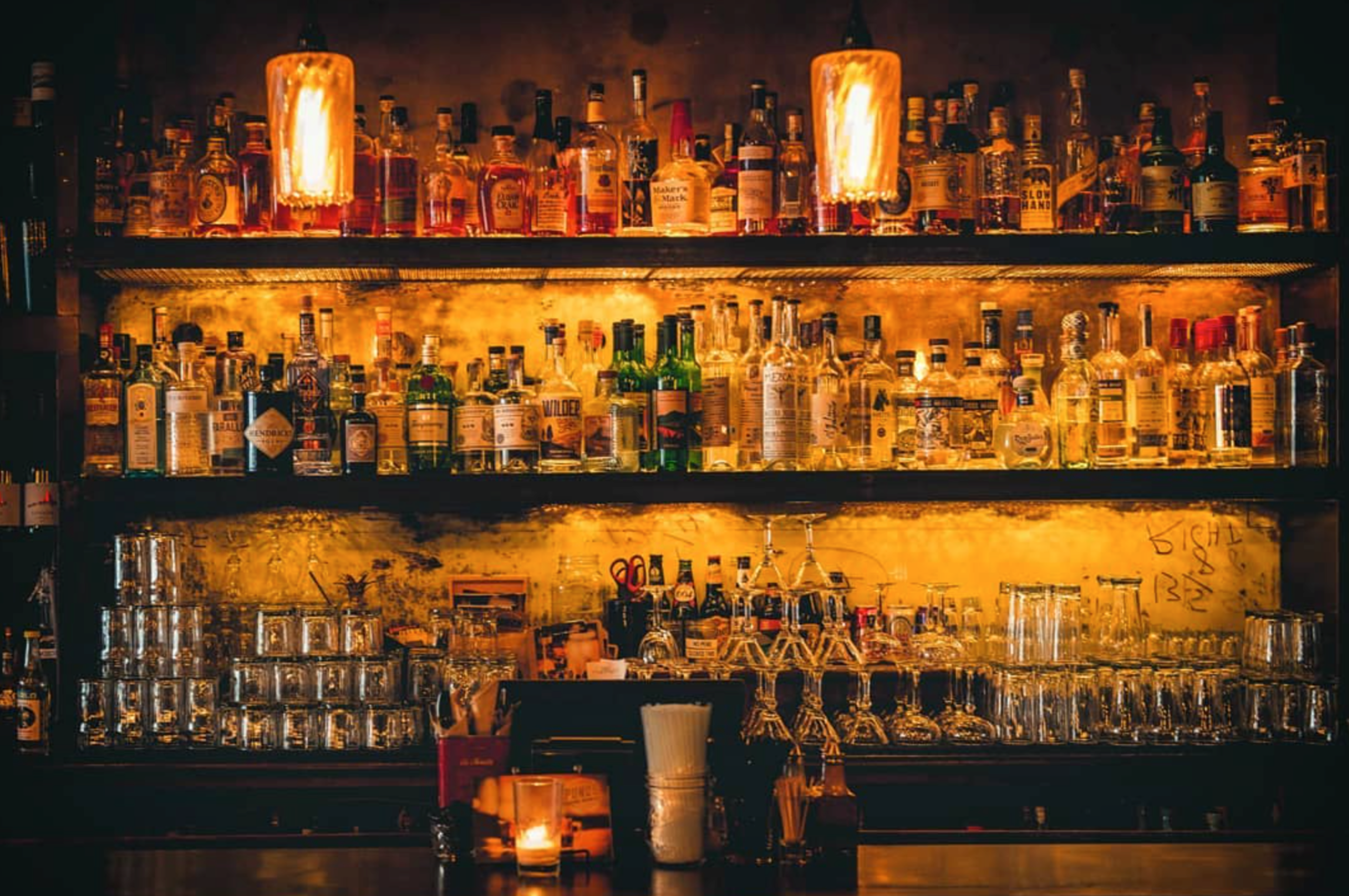 Shelves with various liquor bottles and stacked glasses are illuminated by two warm pendant lights above a wooden bar counter.