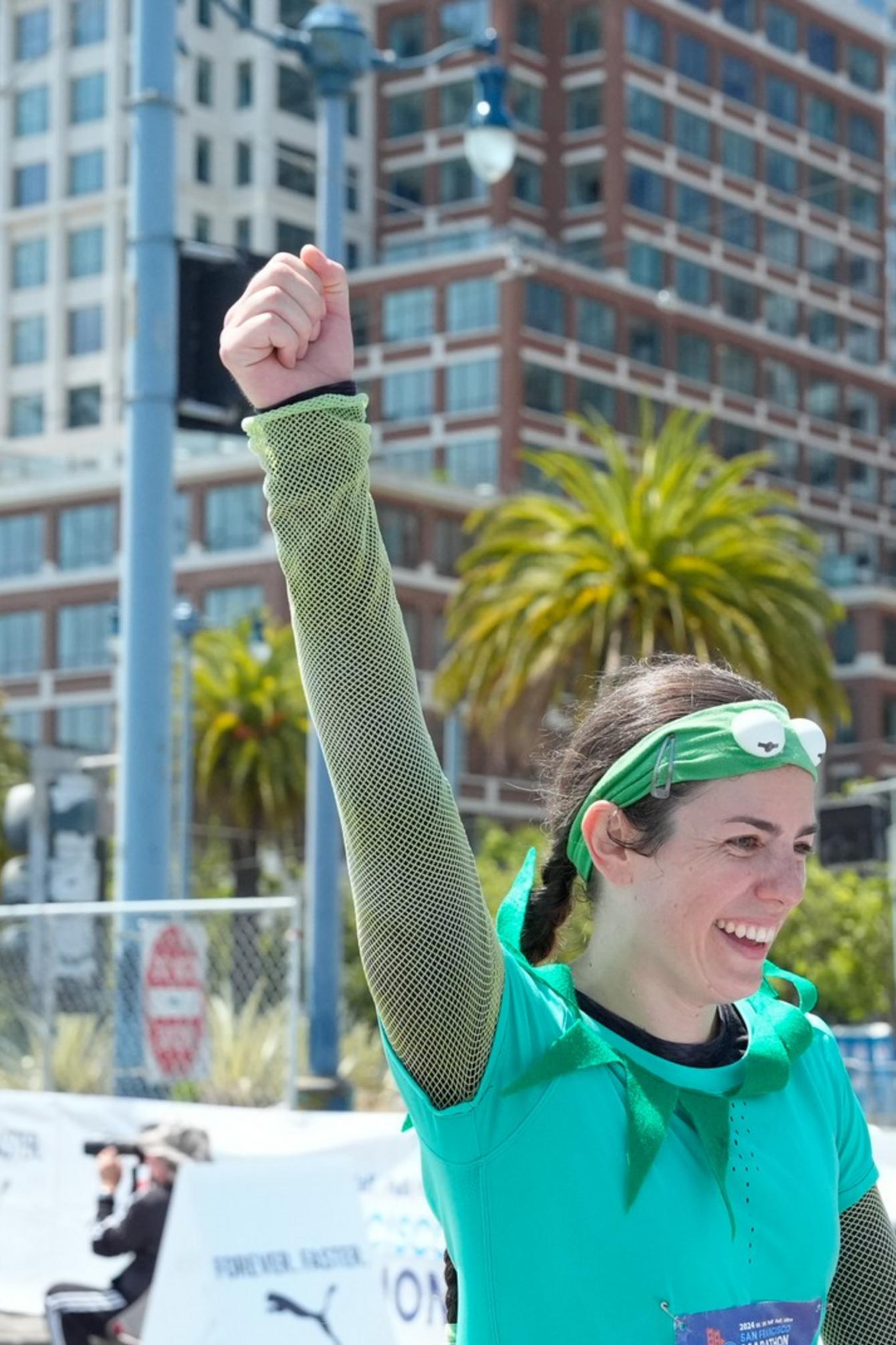 A woman in a teal outfit raises her fist in victory, smiling, with a race bib number &quot;7261.&quot; She's on a city street, with palm trees and tall buildings in the background.