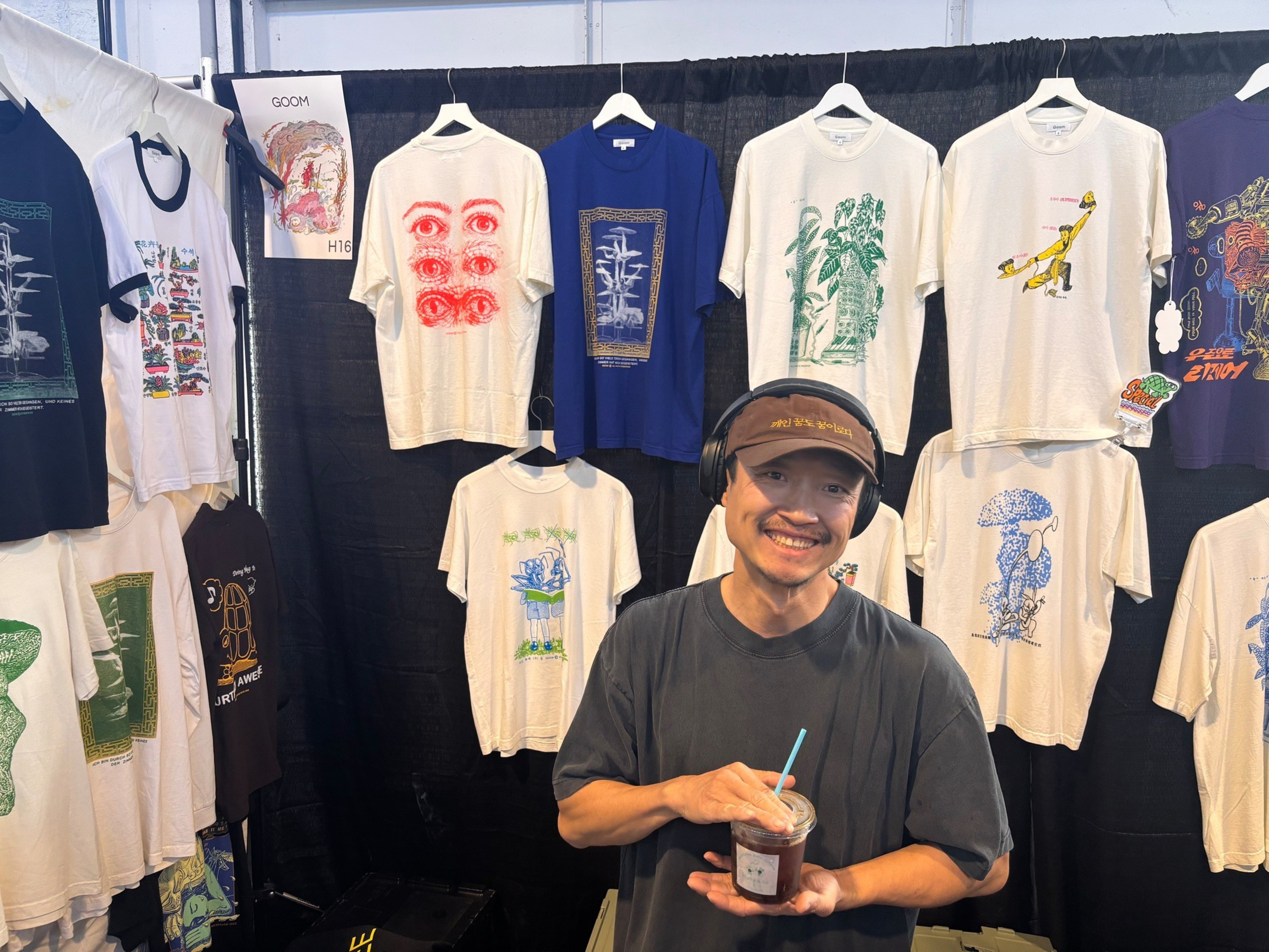 A smiling man wearing a visor and headphones holds a drink in front of a display of various graphic t-shirts hanging on a black backdrop.