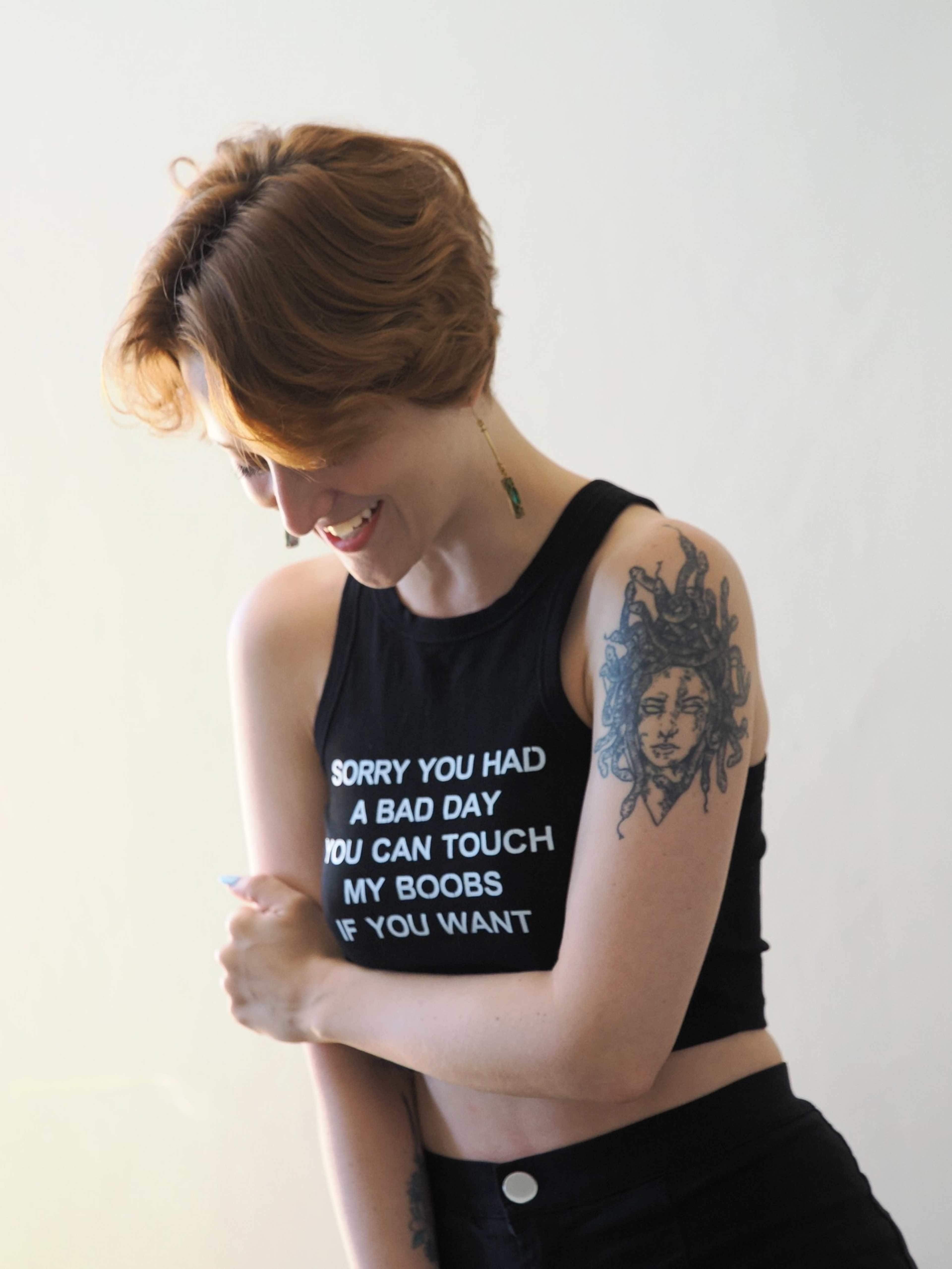 A smiling person with short brown hair wears a black crop top with white text and a detailed tattoo of Medusa on their upper arm.