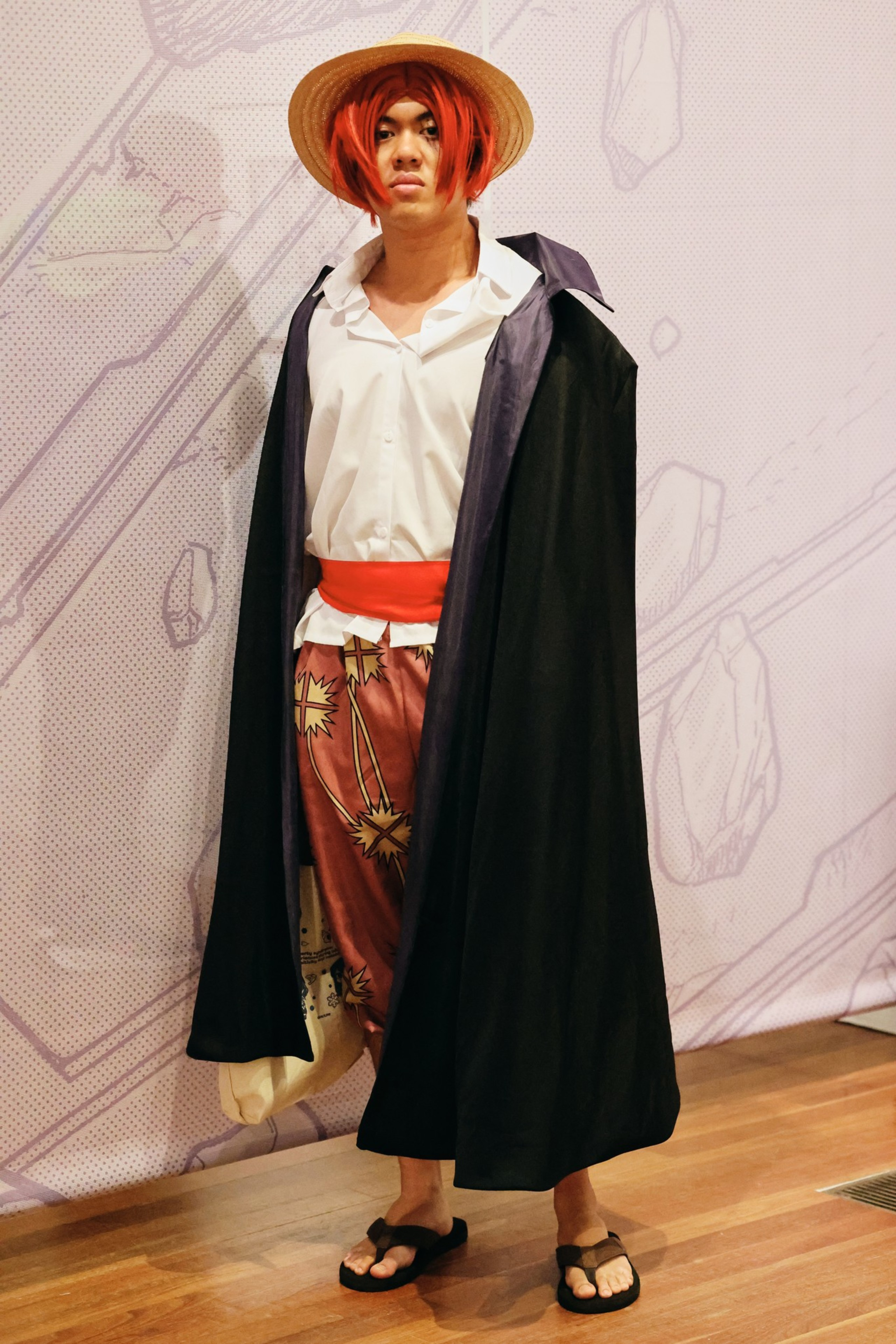 A person wears a straw hat, red wig, white shirt, red sash, patterned pants, black cloak, and sandals, standing against a light patterned wall.