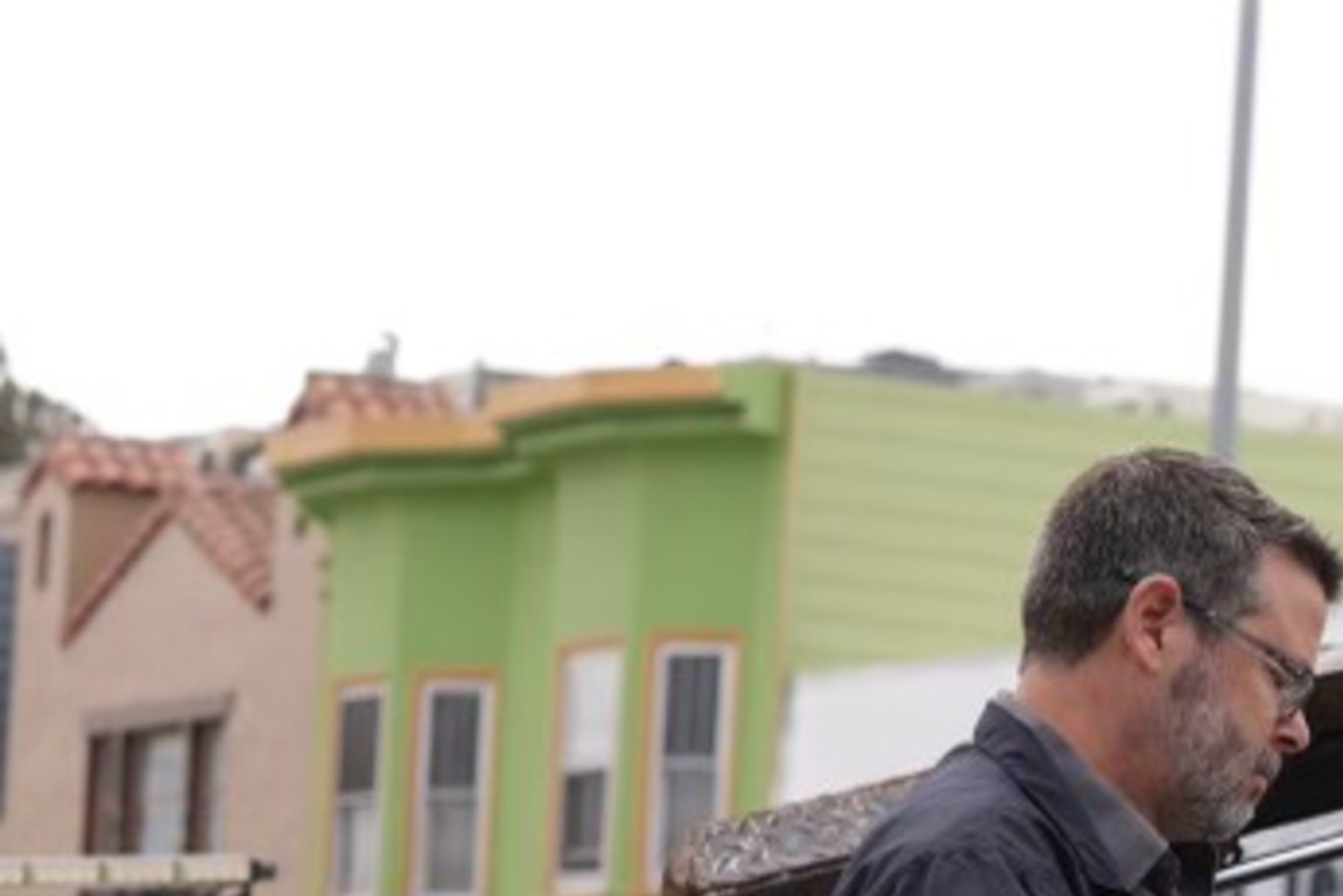 An FBI agent wearing gloves examines items in the open back of a black "Specialty Towing" truck parked on a street next to white and green buildings.