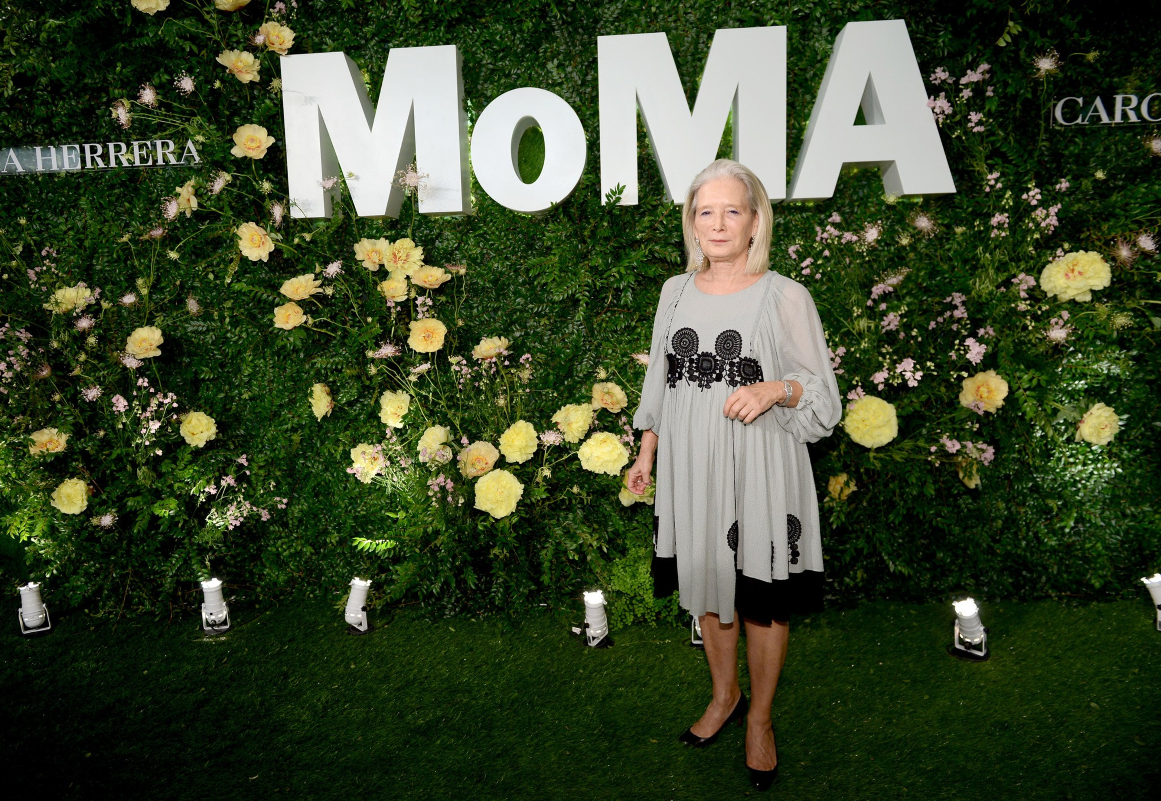 A woman stands in front of a lush green backdrop with &quot;MoMA&quot; in large white letters, surrounded by yellow flowers and smaller text. She wears a grey dress.