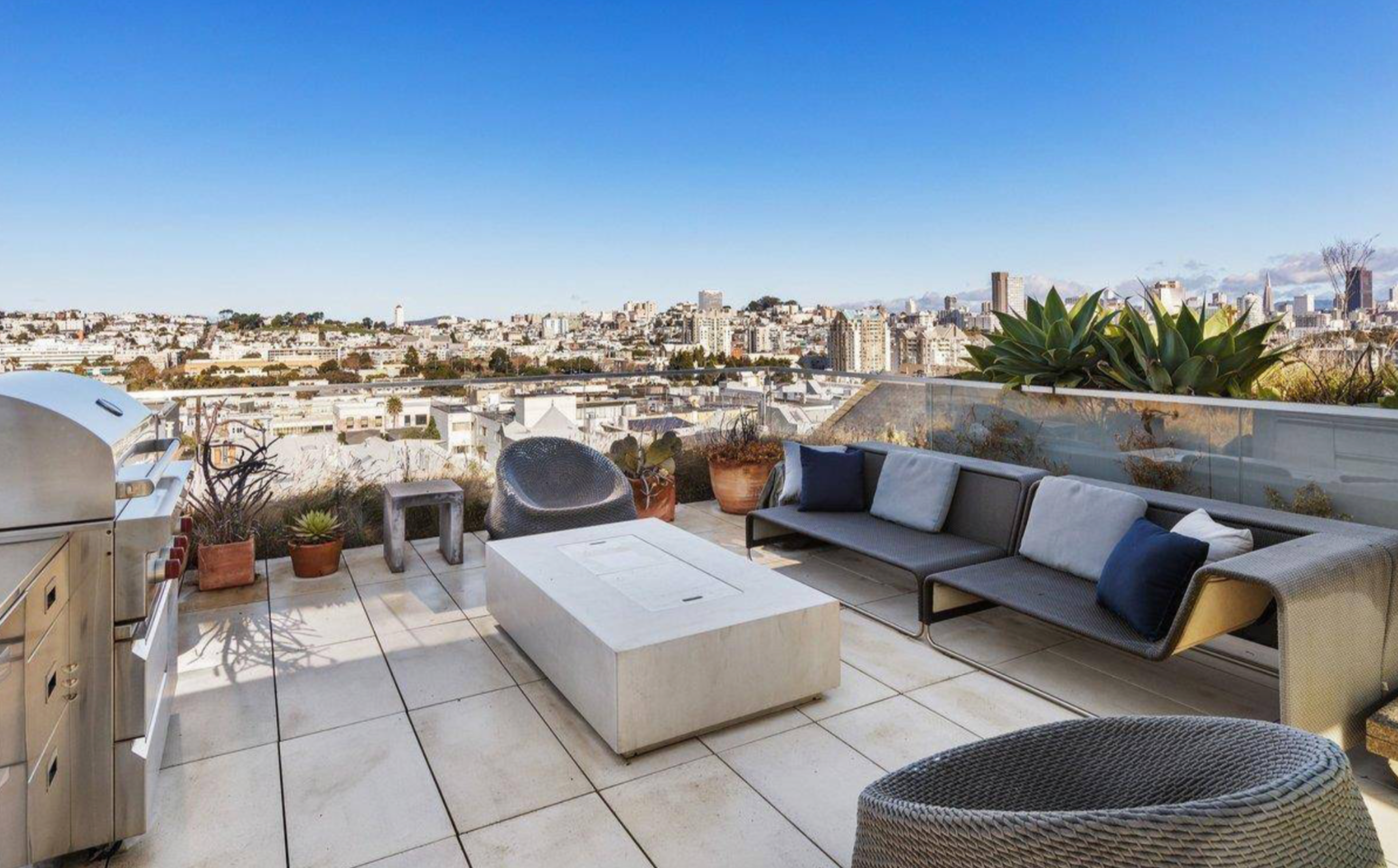 A rooftop patio features a gray sectional sofa, round chairs, a rectangular coffee table, potted plants, and a stainless steel grill with a cityscape backdrop.
