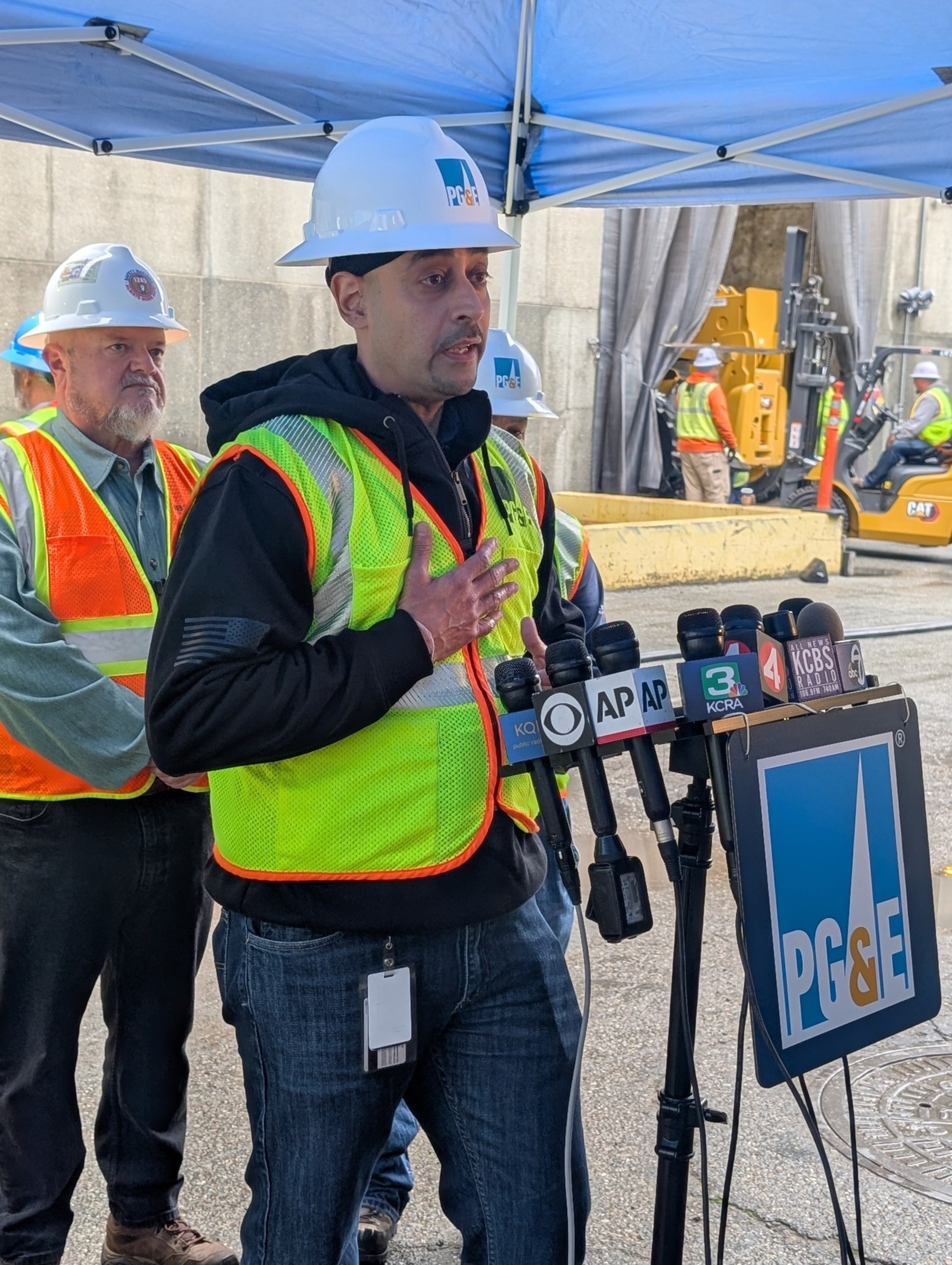 A man wearing a PG&E hard hat and safety vest speaks at a microphone stand with various news outlet logos, while others in safety gear stand behind him.