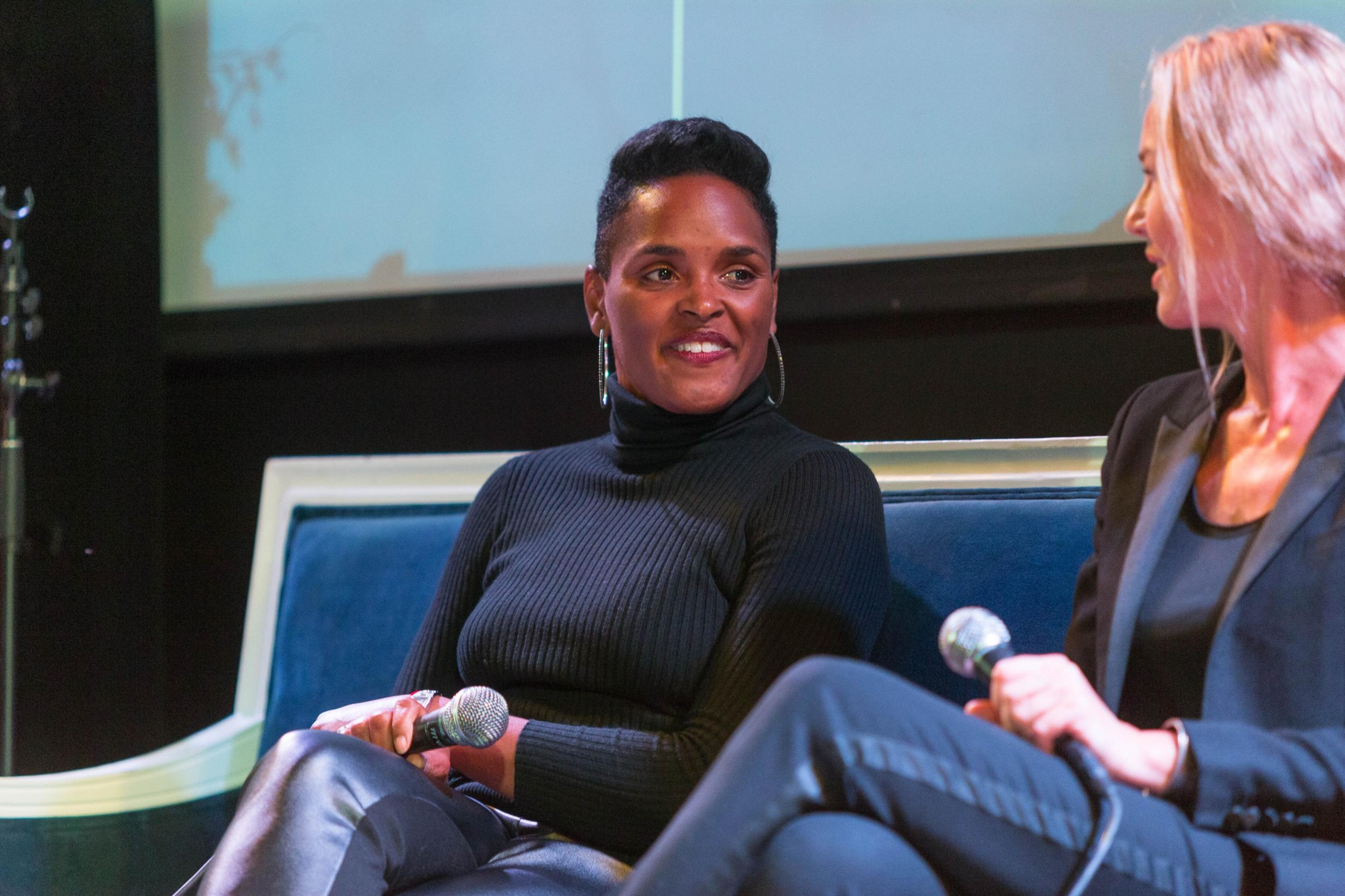 Two people are seated on a stage, each holding a microphone. One wears a black turtleneck, and the other is in a dark blazer. They appear to be in conversation.