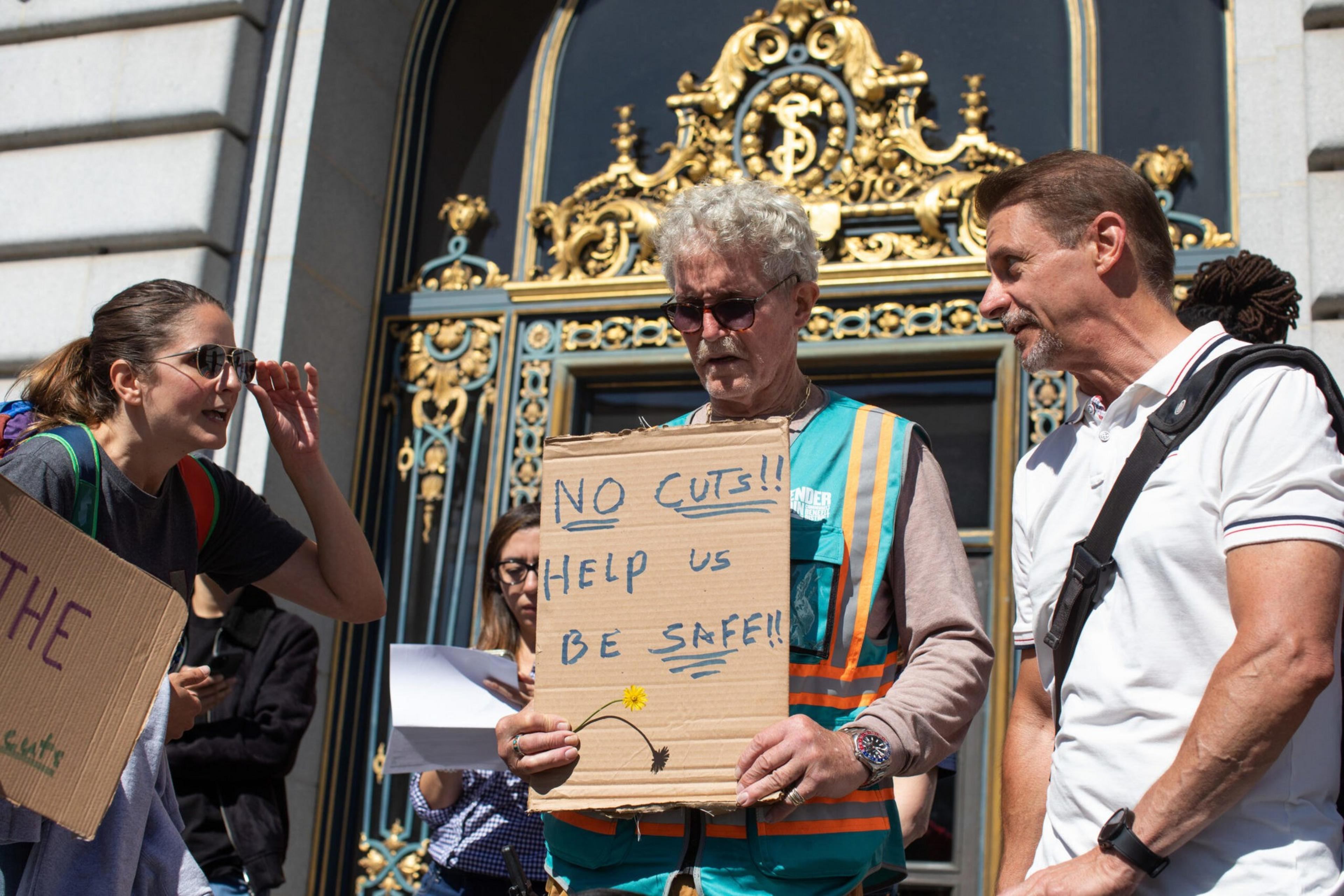 A man in a safety vest holds a cardboard sign reading "NO CUTS!! HELP US BE SAFE!!" while people around him engage in conversation outside a building with ornate gold decorations.
