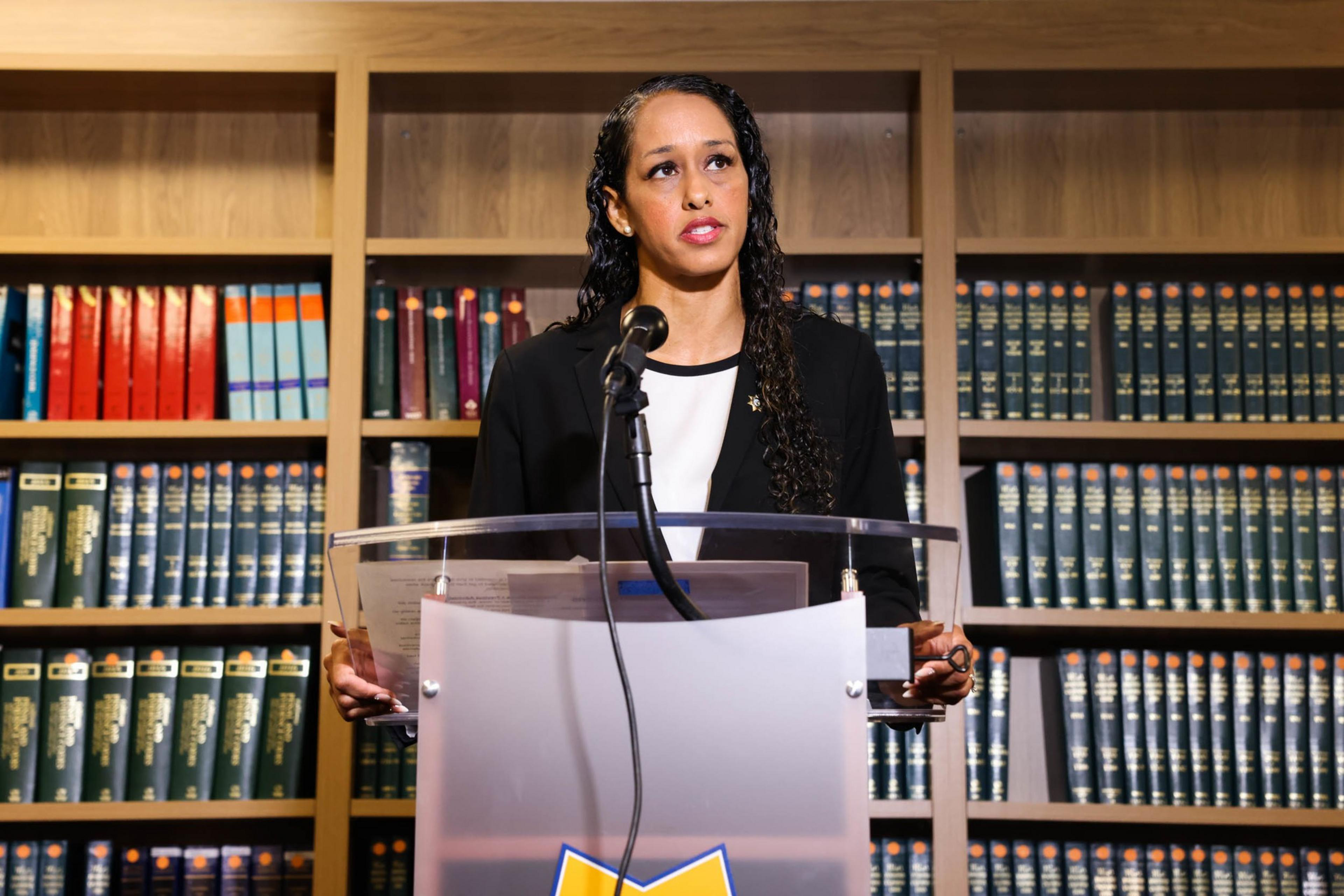 A woman with long curly hair stands behind a clear podium, speaking into a microphone with bookshelves filled with legal books in the background.