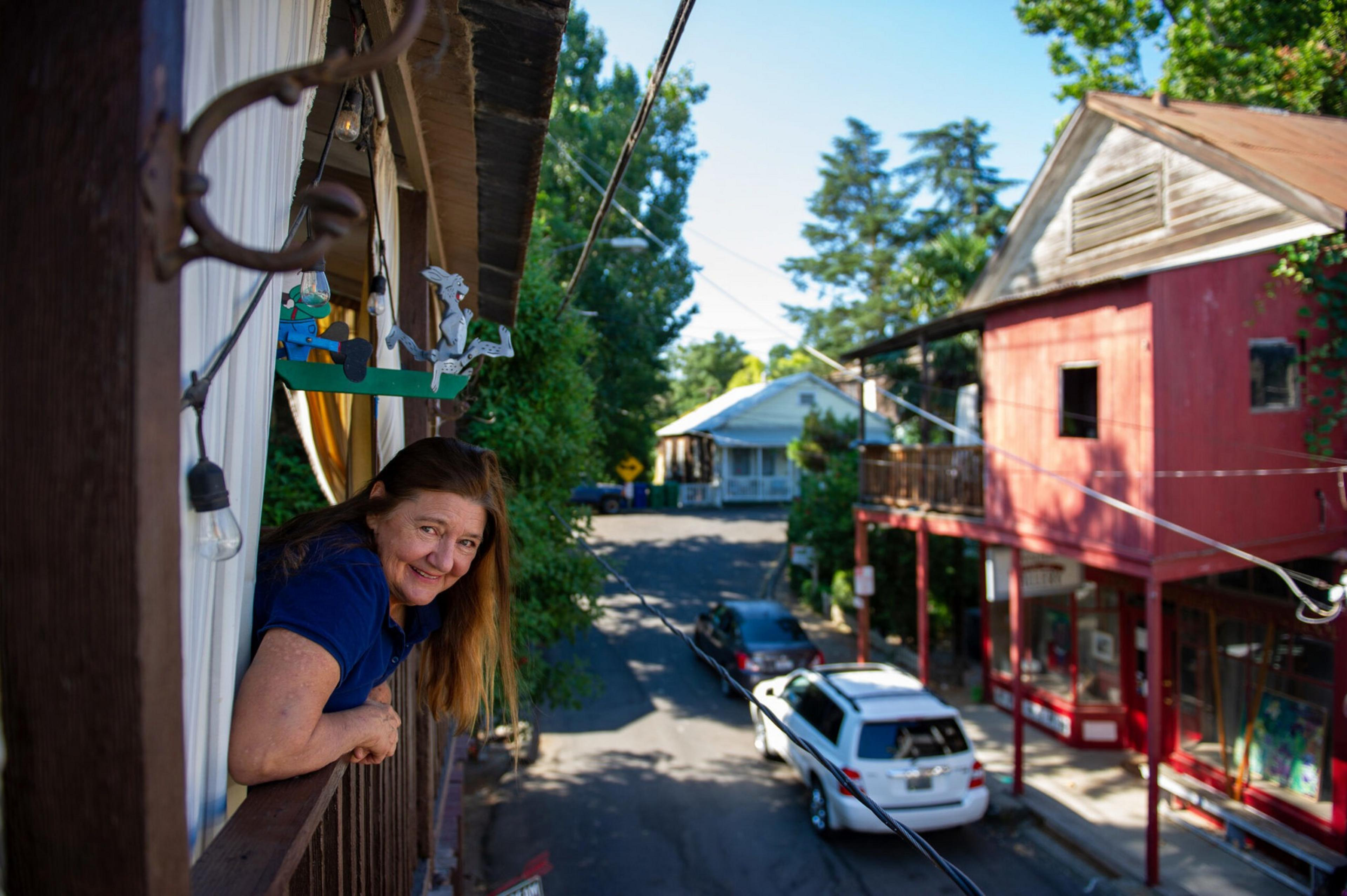A woman leans on a balcony railing, smiling, above a street with parked cars and old buildings under clear blue sky.