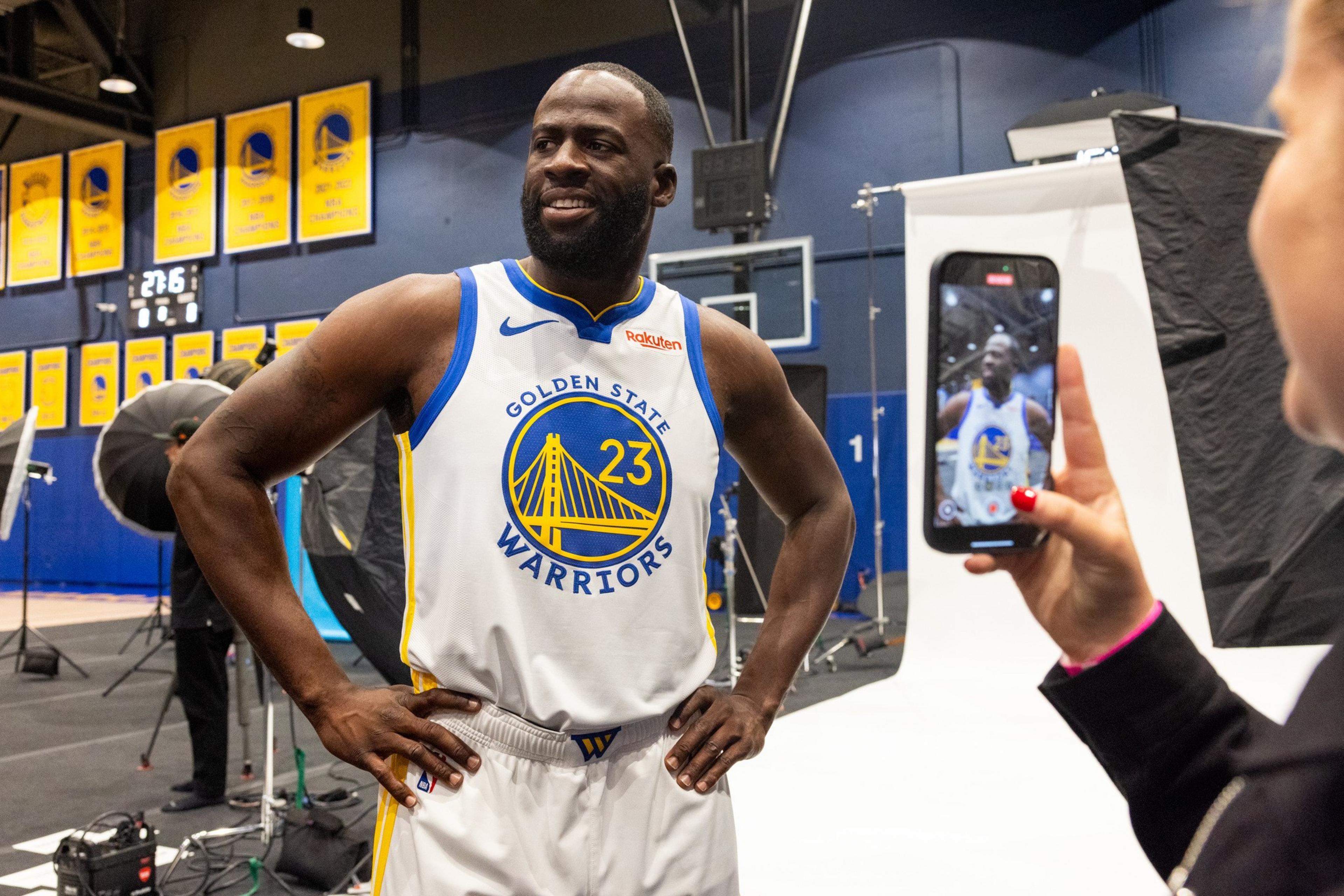 A man wearing a Golden State Warriors basketball jersey with number 23 poses hands-on-hips as someone takes his photo with a smartphone.