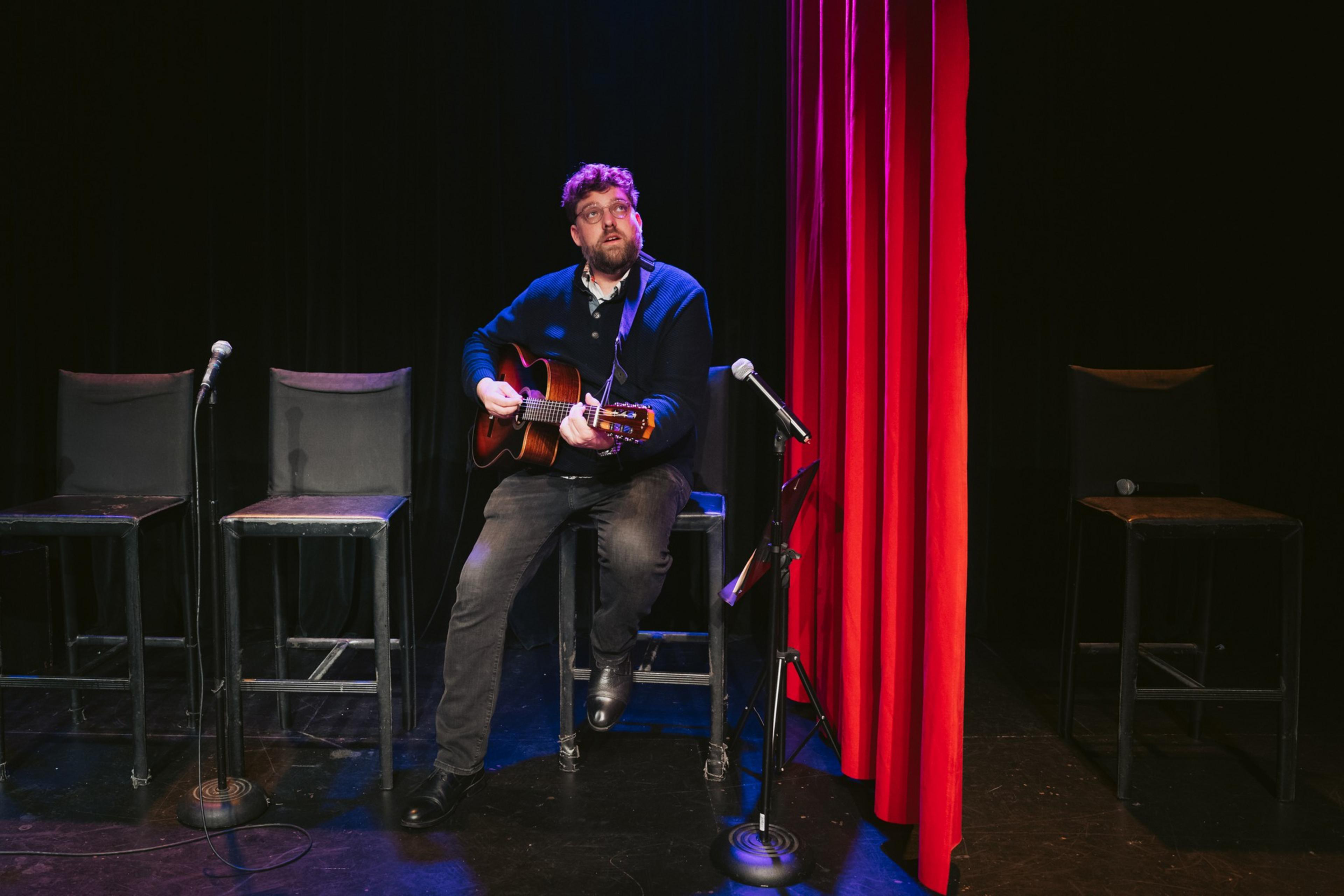 A man sits on a stool playing an acoustic guitar on a dimly lit stage with red curtains and three empty stools nearby.