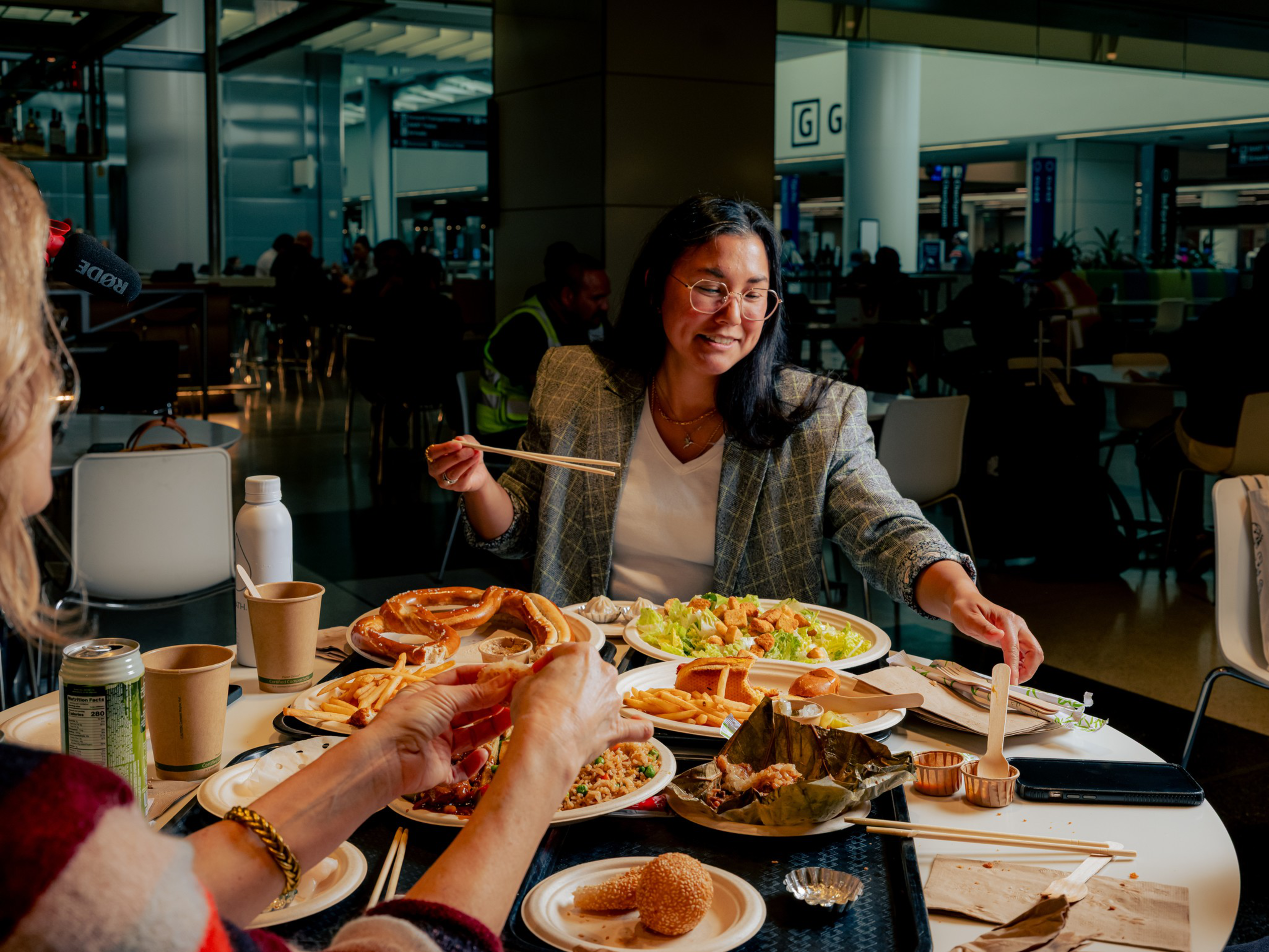 A woman with glasses wearing a plaid blazer uses chopsticks to eat from plates of various foods including salad, fried rice, and rolls at a busy dining table.