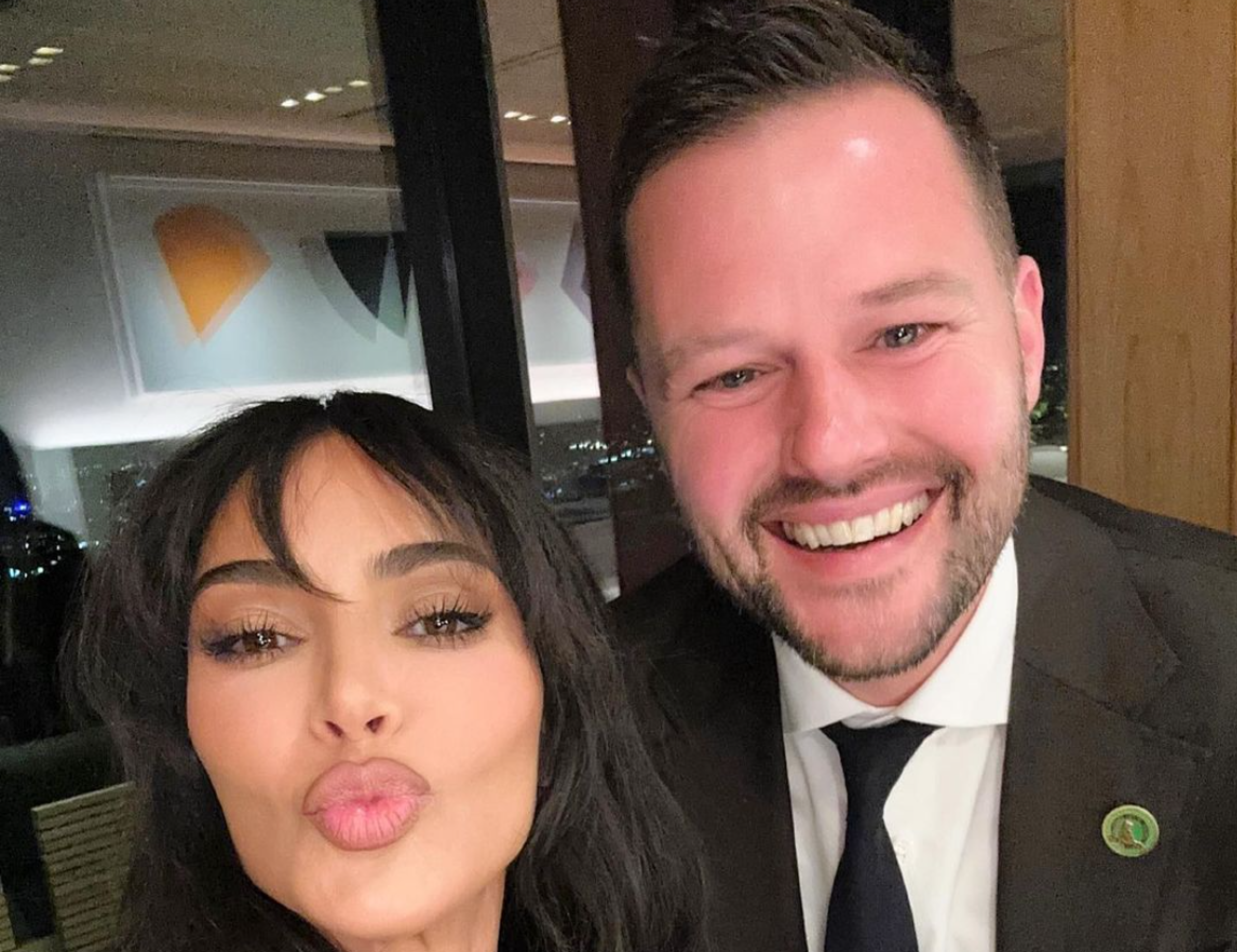 A woman with long dark hair puckers her lips while a man with a beard and suit smiles broadly behind her.