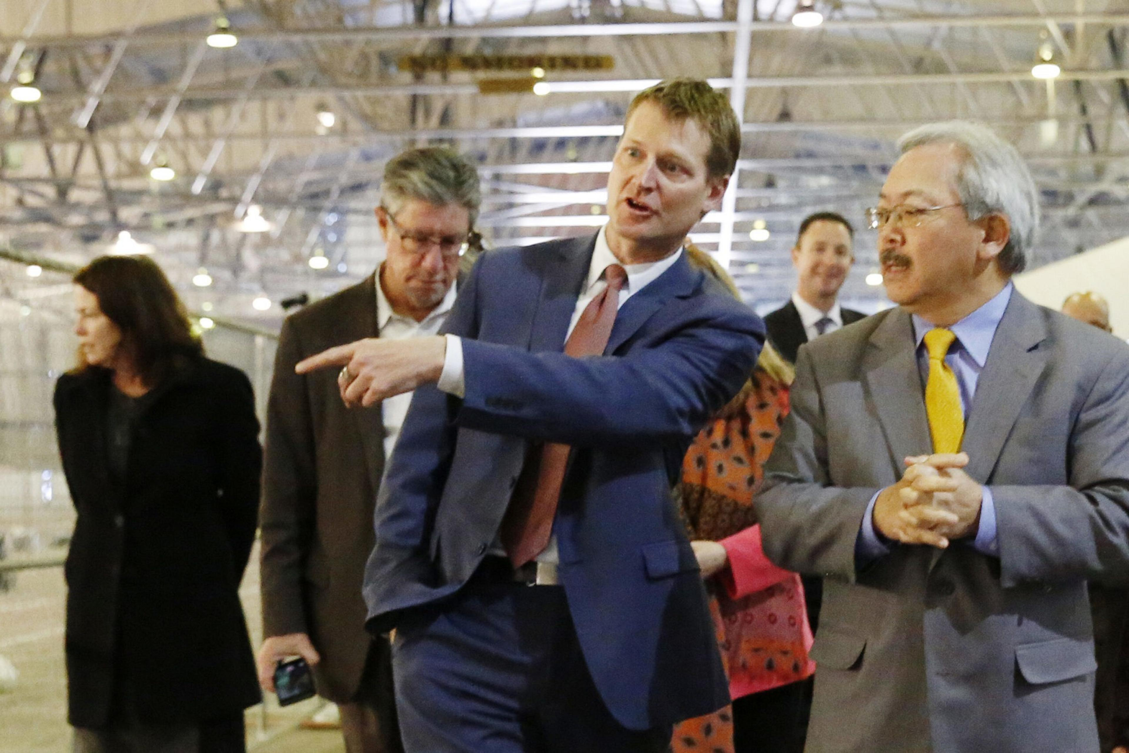 A man in a blue suit points while talking to another man in a gray suit with a yellow tie, as three other people follow behind them.