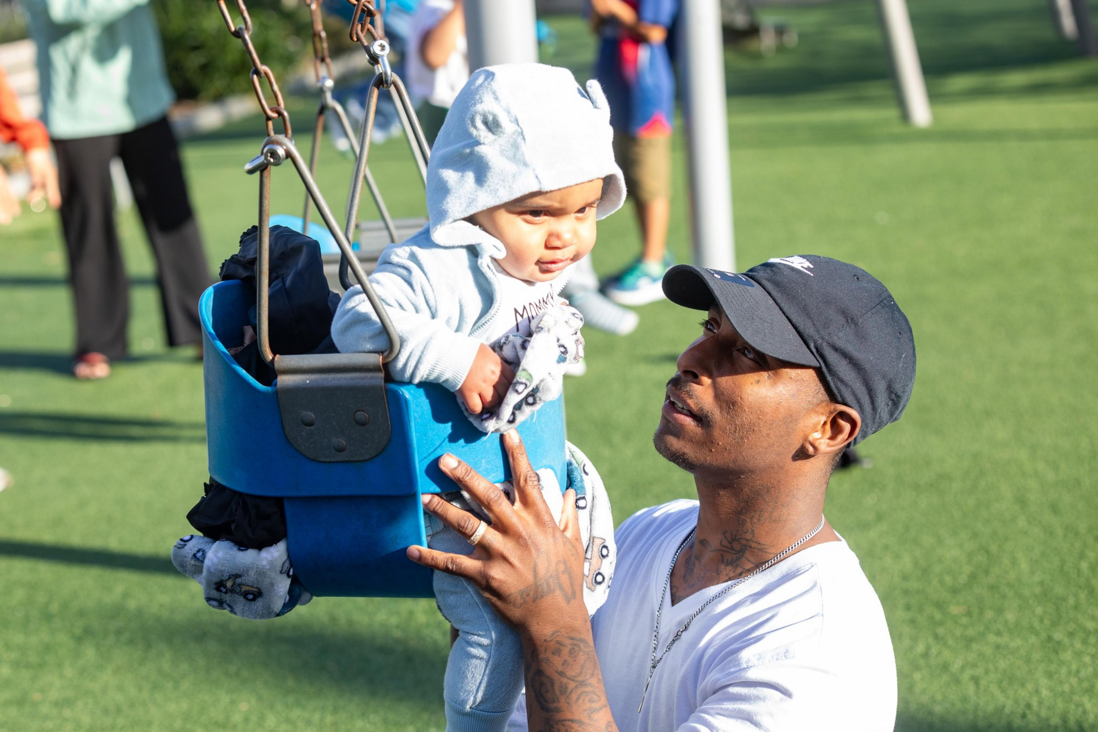 A man pushes a baby in a blue swing. The baby, wearing a hoodie, smiles while the man holds the swing, looking at the baby with care.