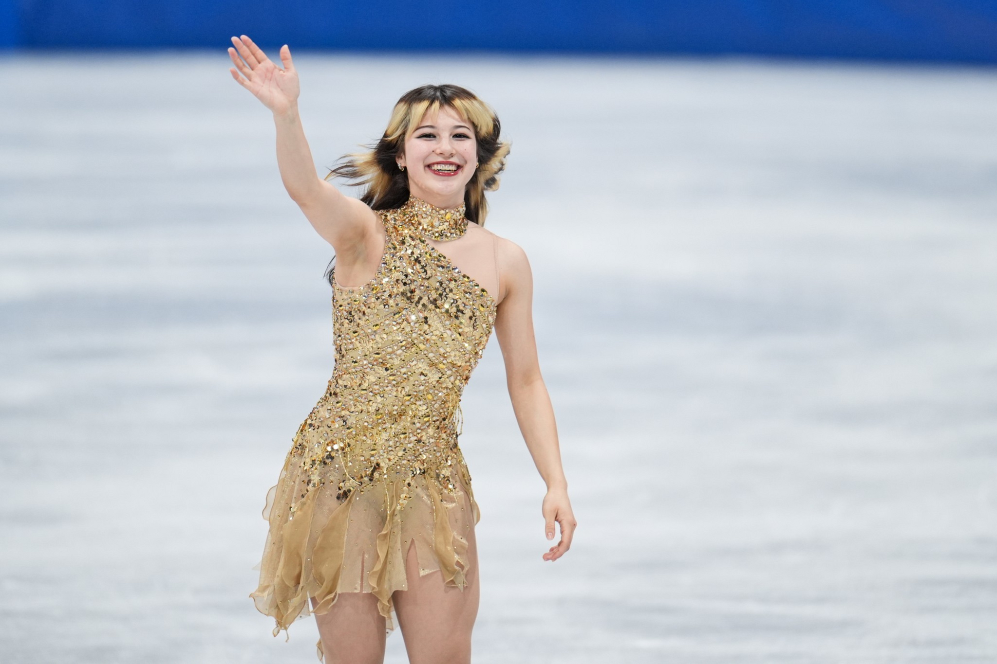 A female figure skater in a sparkling gold dress smiles and waves with her right hand while standing on the ice rink.