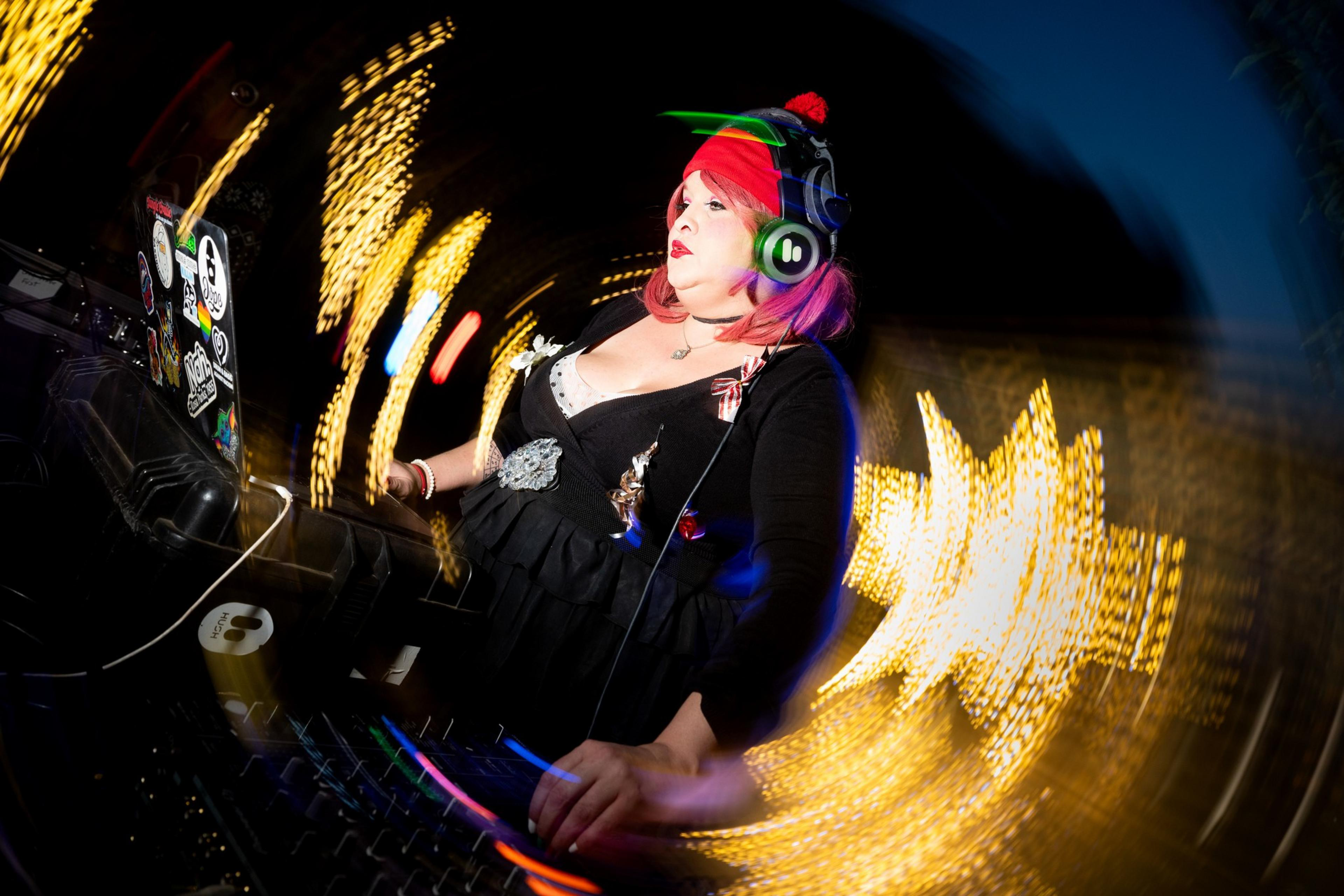 A DJ with pink hair, wearing headphones and a festive outfit, is focused on mixing music. The background is a swirl of colorful lights and blurred designs.