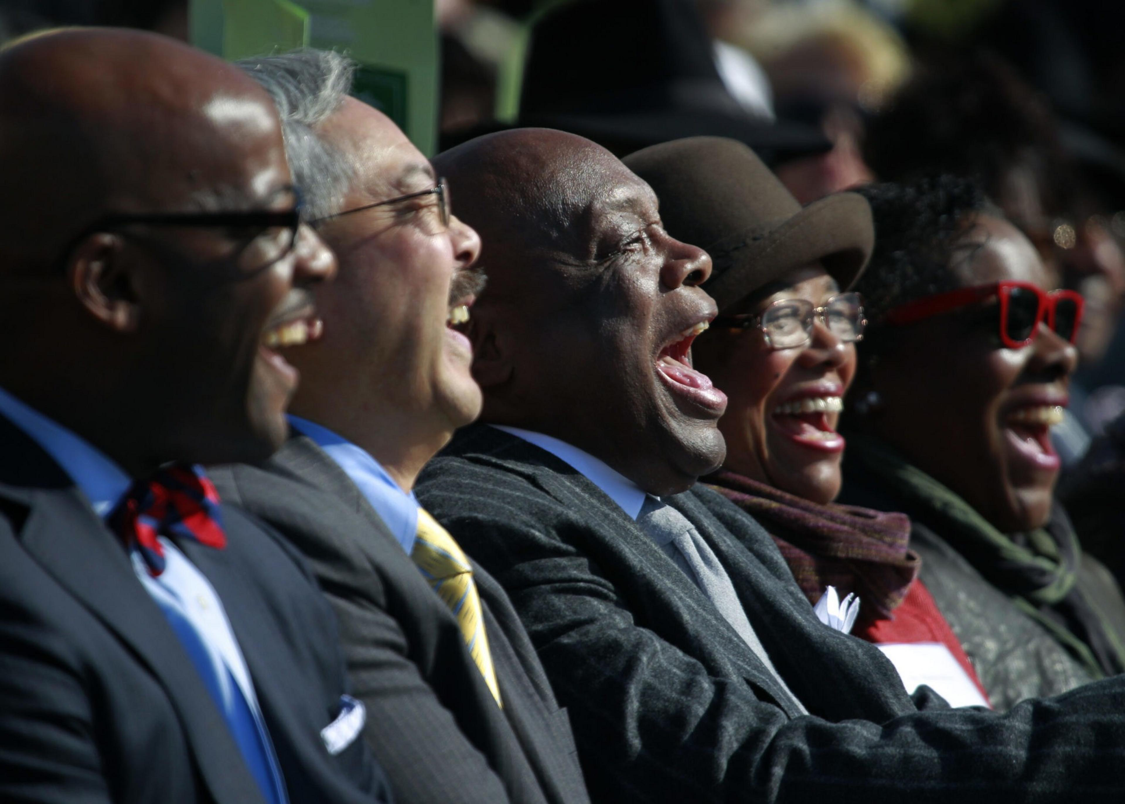 Five people in formal attire sit closely, all laughing heartily, with one wearing bright red glasses and another in a brown hat.