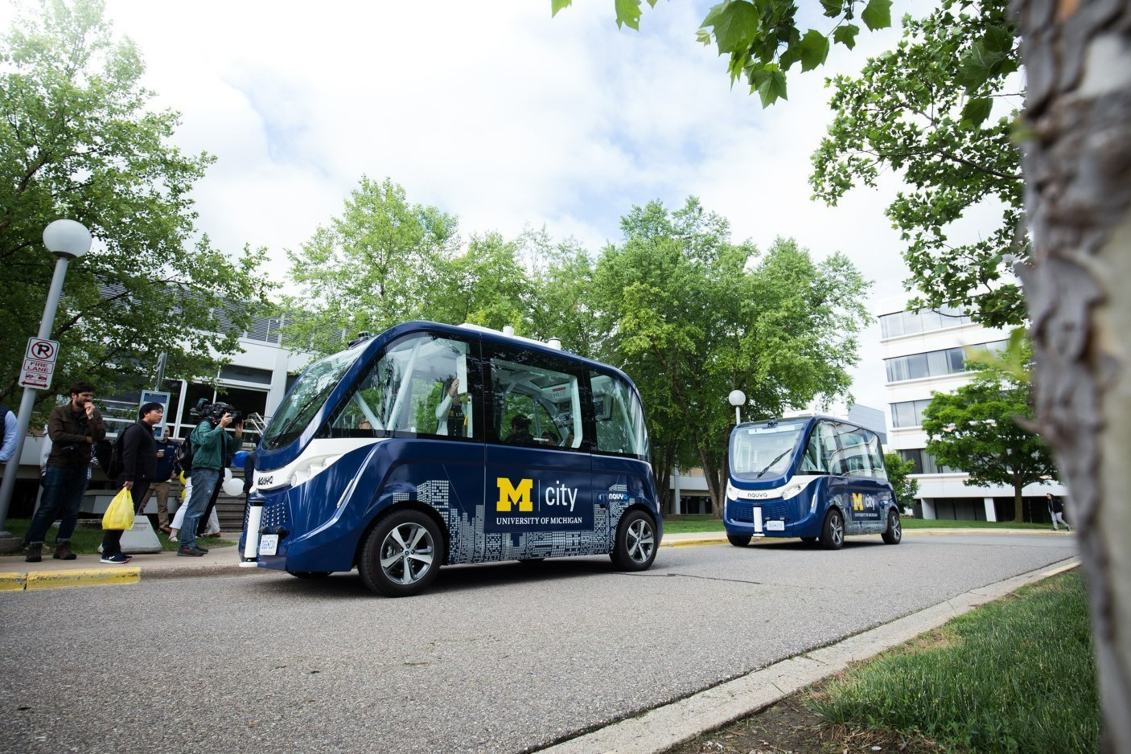 Two small, blue autonomous shuttle vehicles labeled "M city University of Michigan" drive on a campus road with trees and buildings nearby.