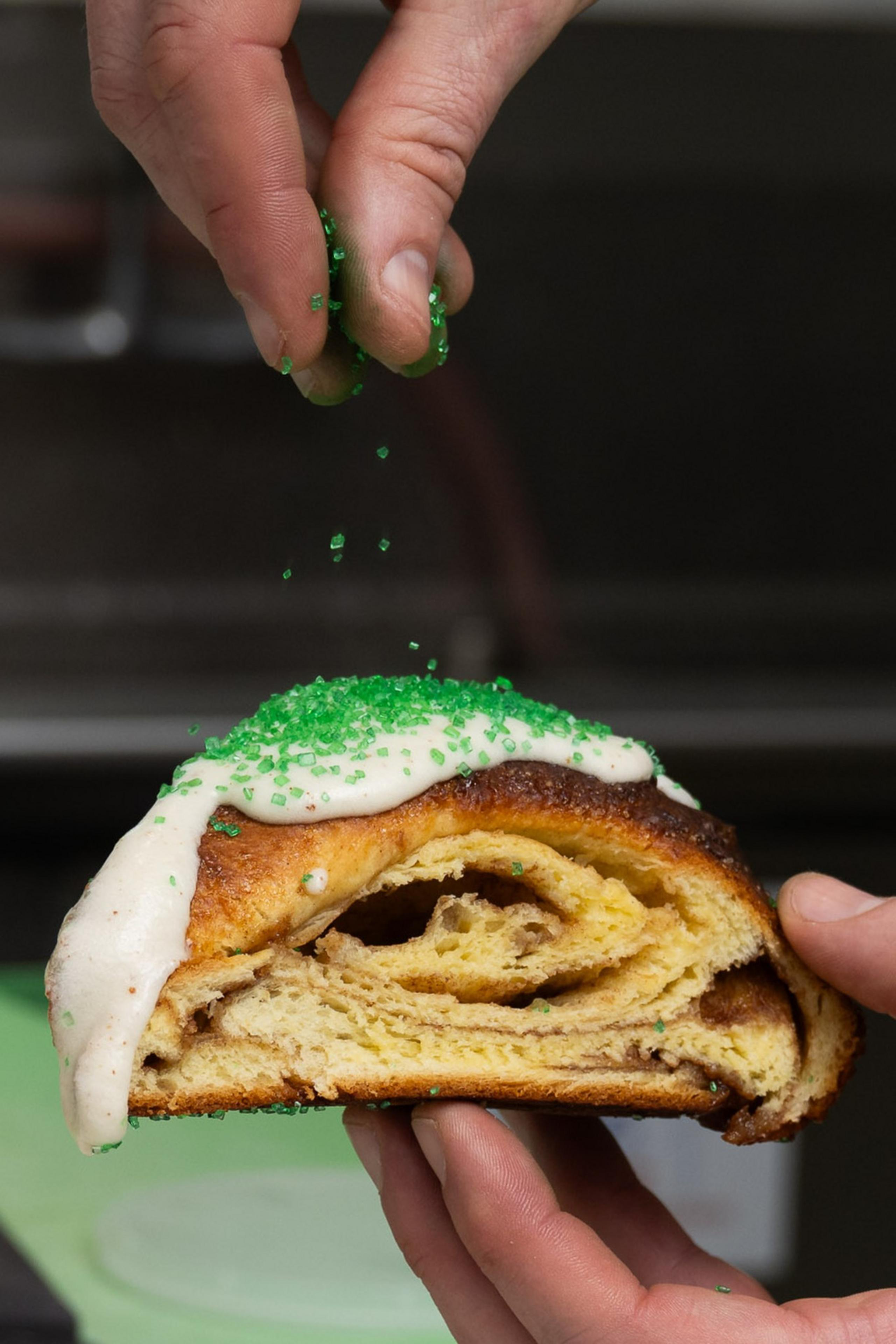 A hand holds a slice of cinnamon bread with icing, while another hand sprinkles green sugar on top.