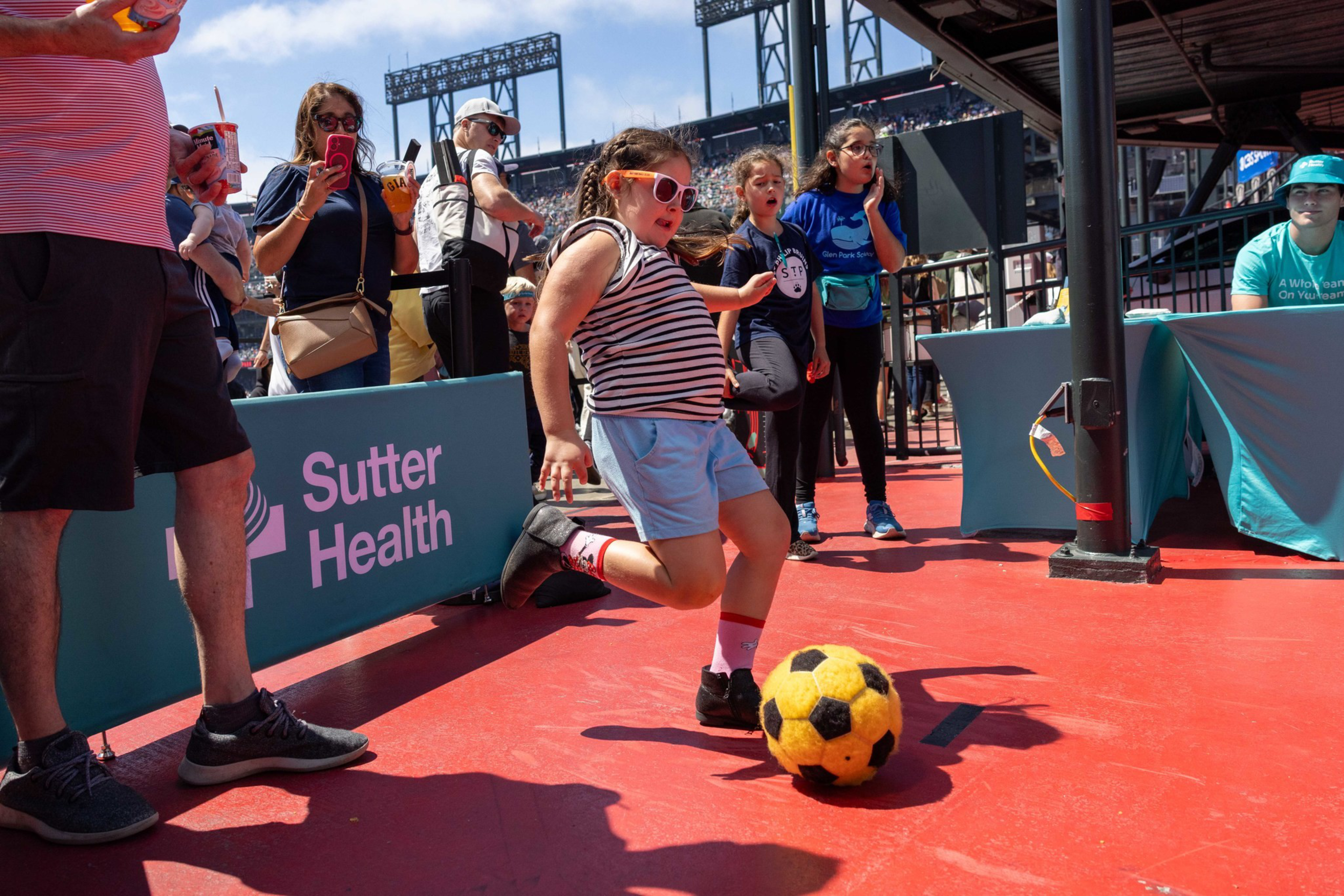 A young girl wearing sunglasses and striped shirt kicks a yellow soccer ball on a red surface while people watch and take photos nearby.