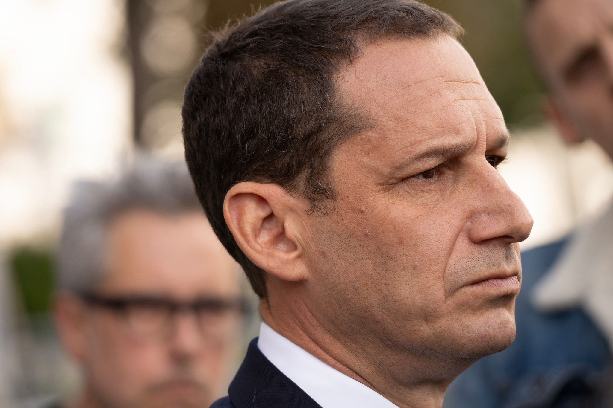 A man with short dark hair and a serious expression faces right, with two blurred figures in the background.