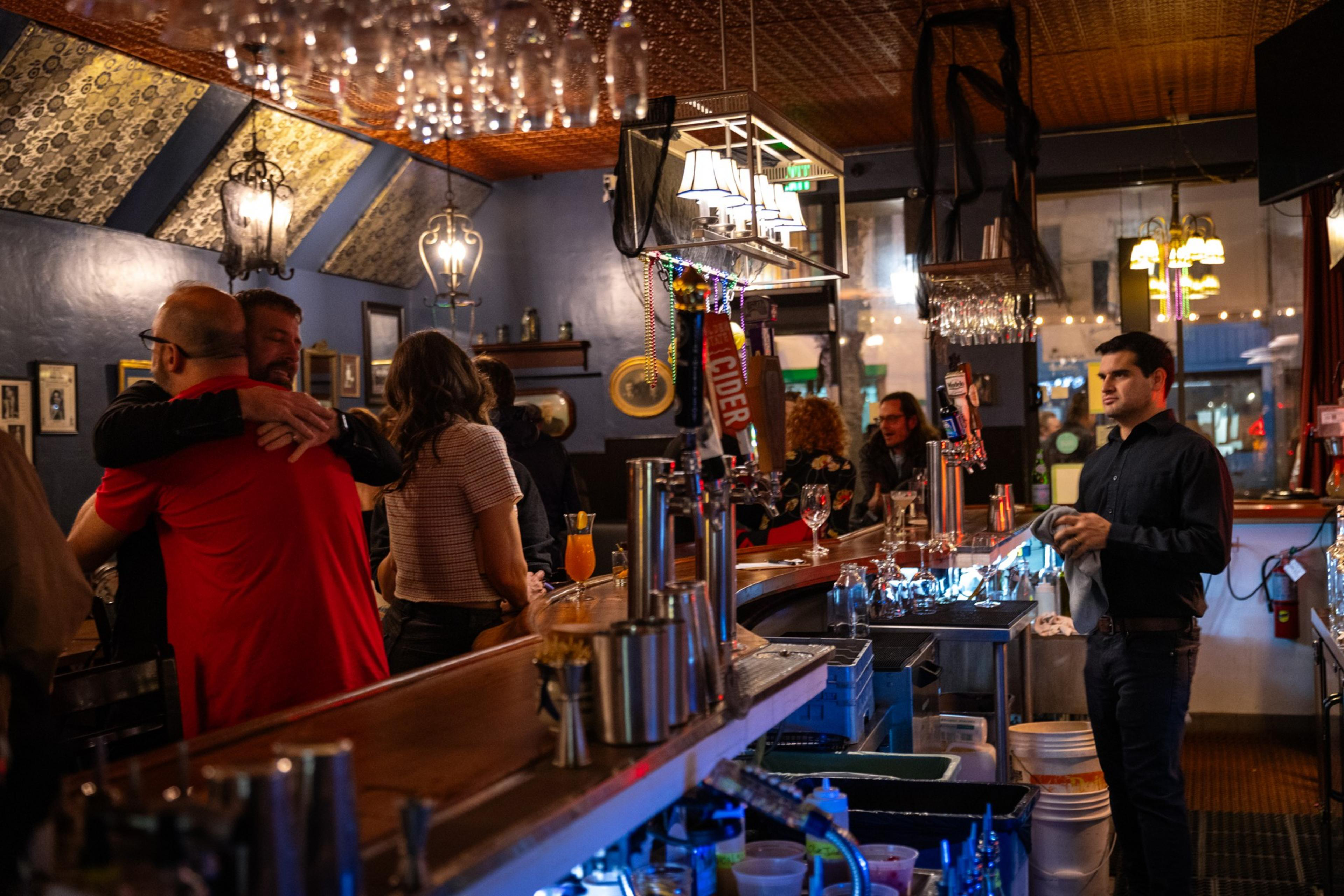 A group of people stand around talking while 2 people hug to one another in a bar setting.