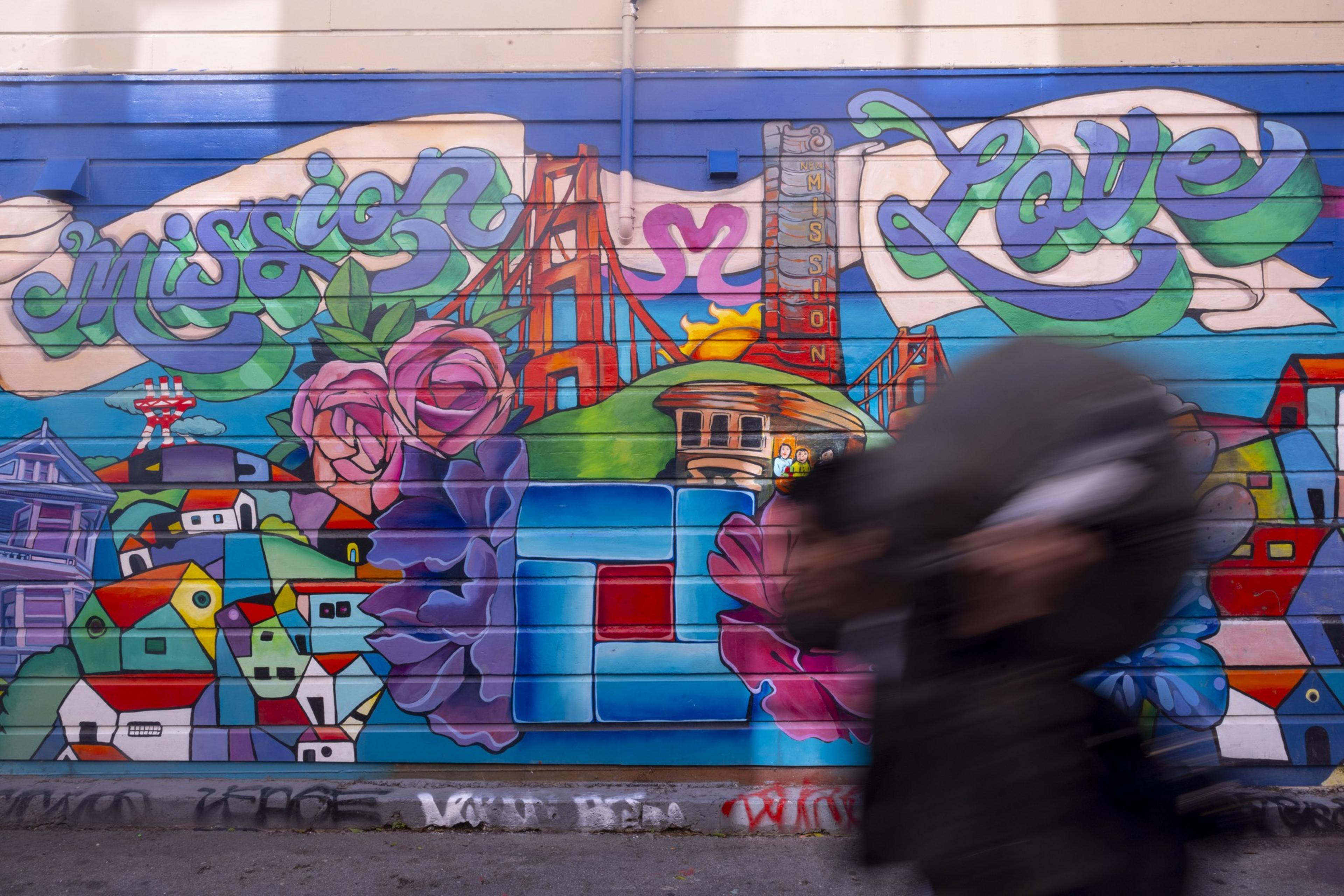 A colorful mural with "Mission Love," featuring the Golden Gate Bridge, roses, houses, and abstract shapes. A blurred person walks by in the foreground.