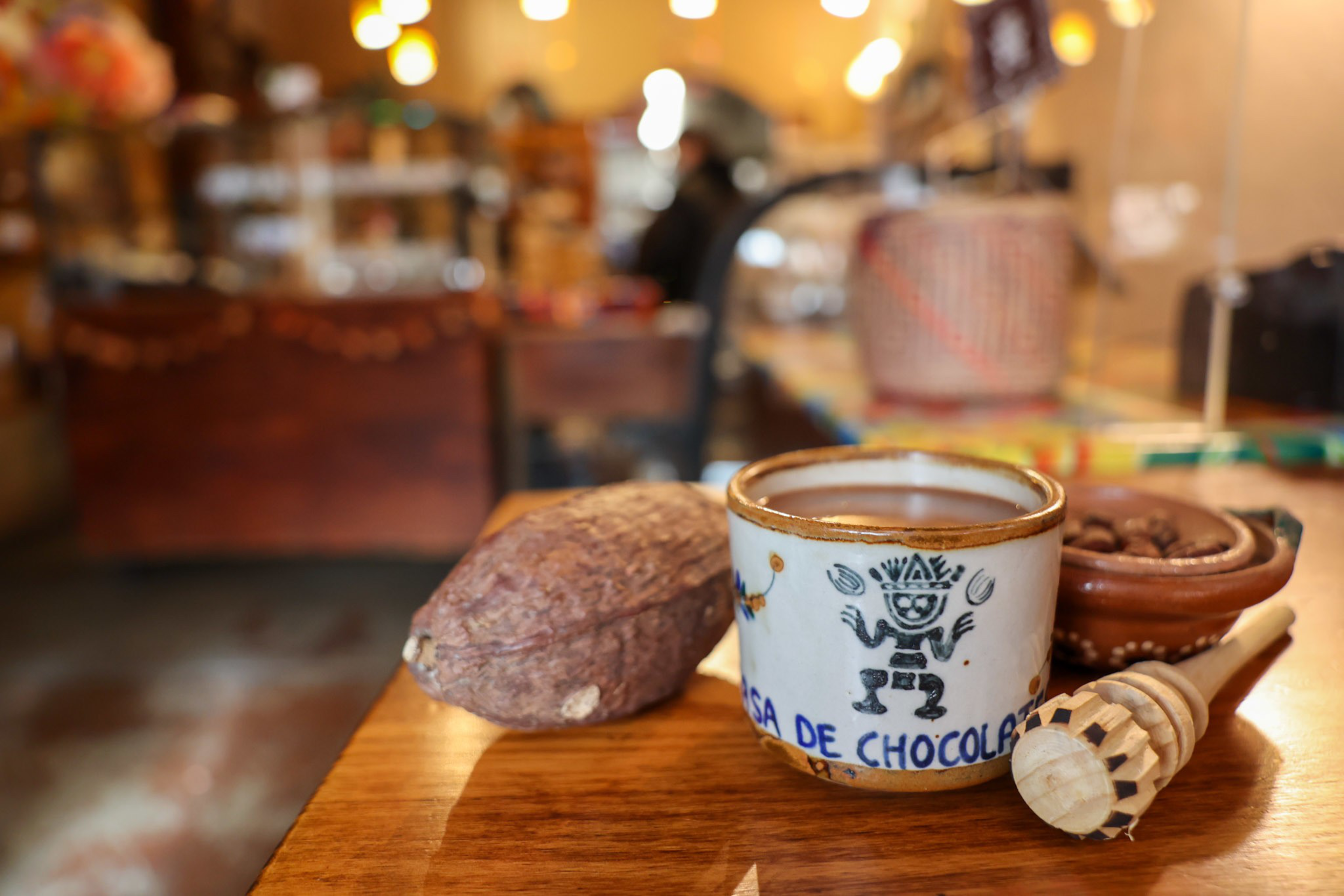 A cup of hot chocolate sits next to a whole cocoa pod, a bowl of cocoa beans, and a wooden honey dipper on a wooden table.