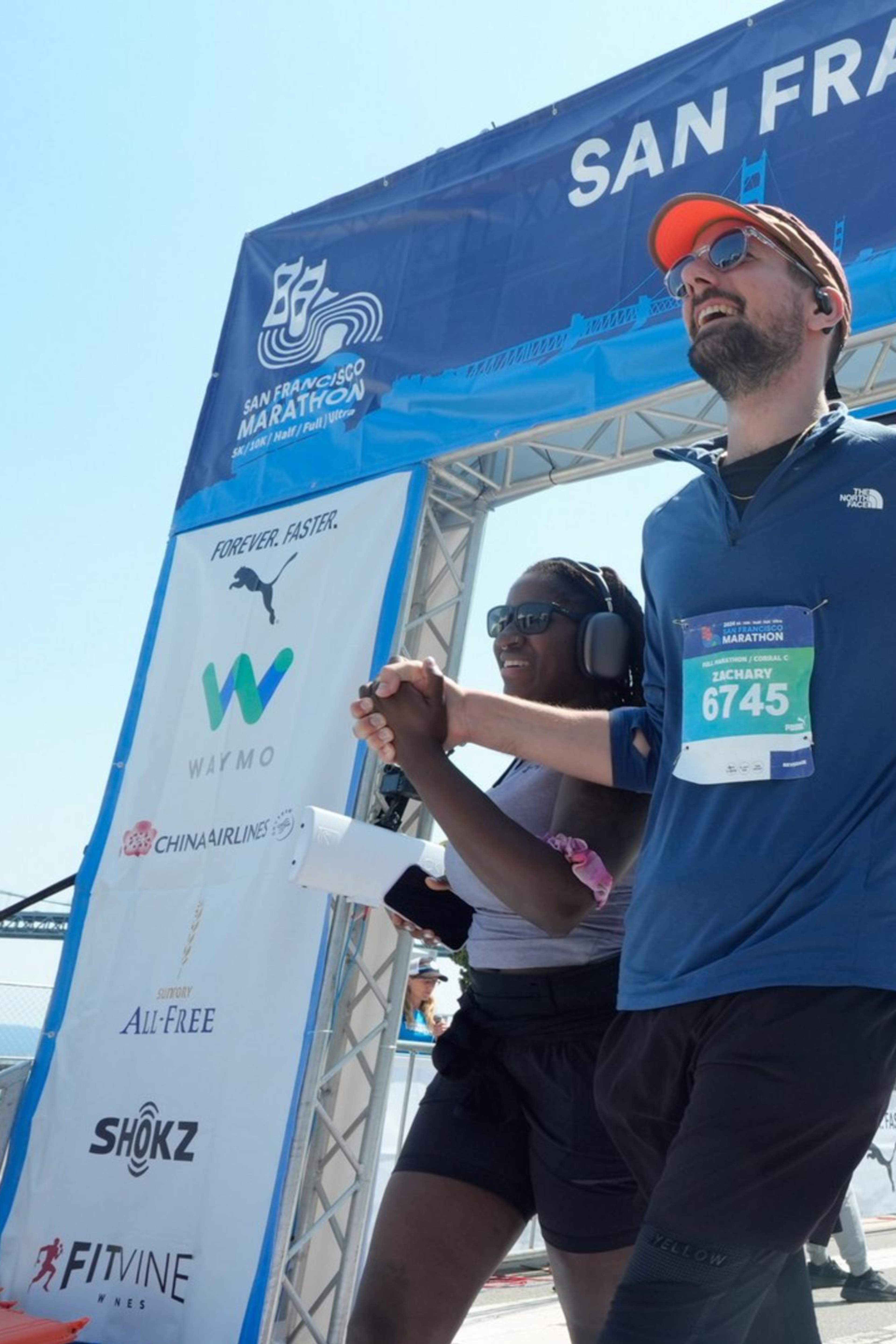 Two runners holding hands cross the finish line of the San Francisco Marathon, smiling. The race clock reads 3:45:29, and a bridge and spectators are visible in the background.