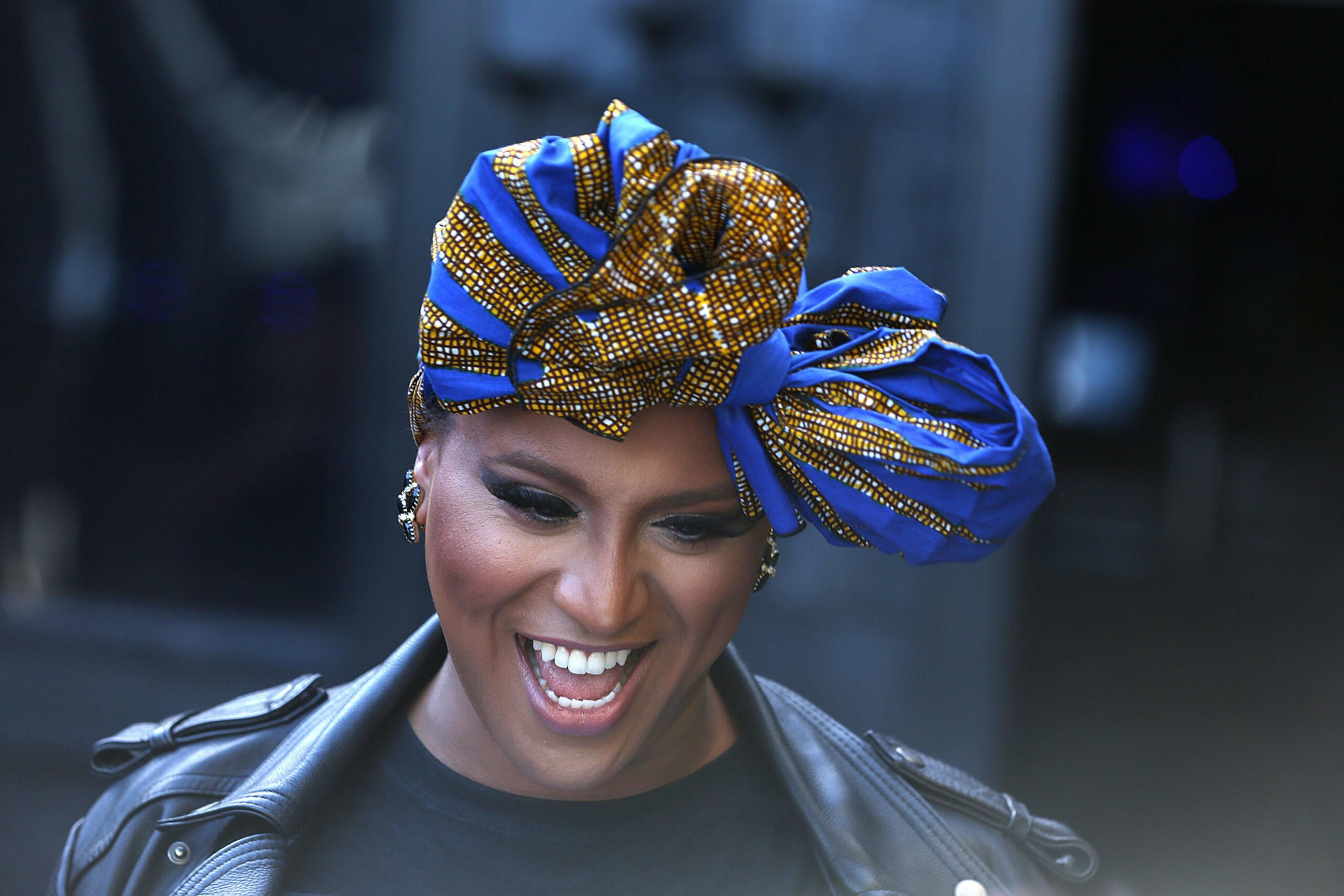 A person with a bright smile wears a blue and gold patterned headwrap, black leather jacket, and statement earrings.