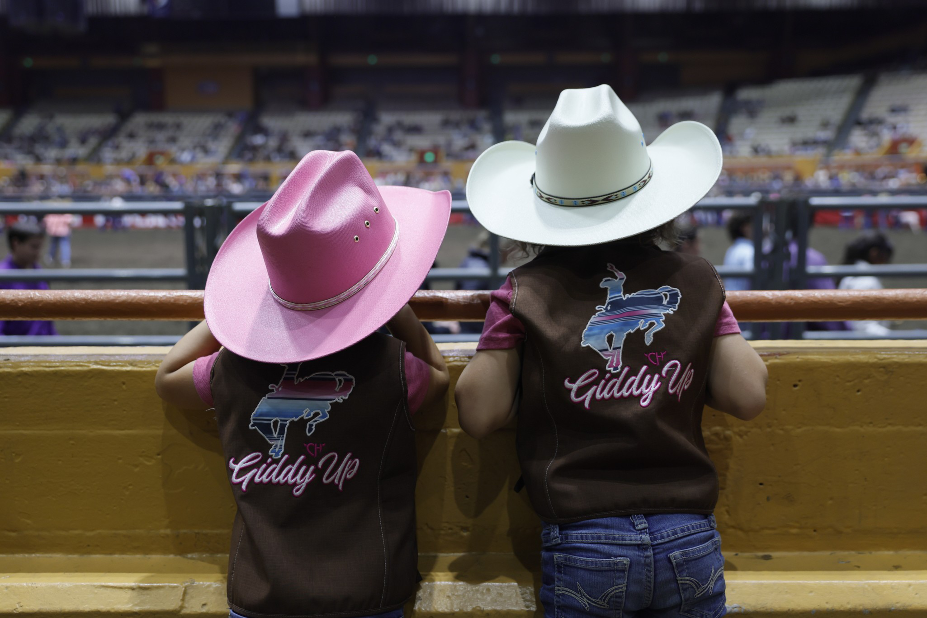 Two children wearing cowboy hats, one pink and one white, lean on a fence watching a rodeo arena inside a large stadium.