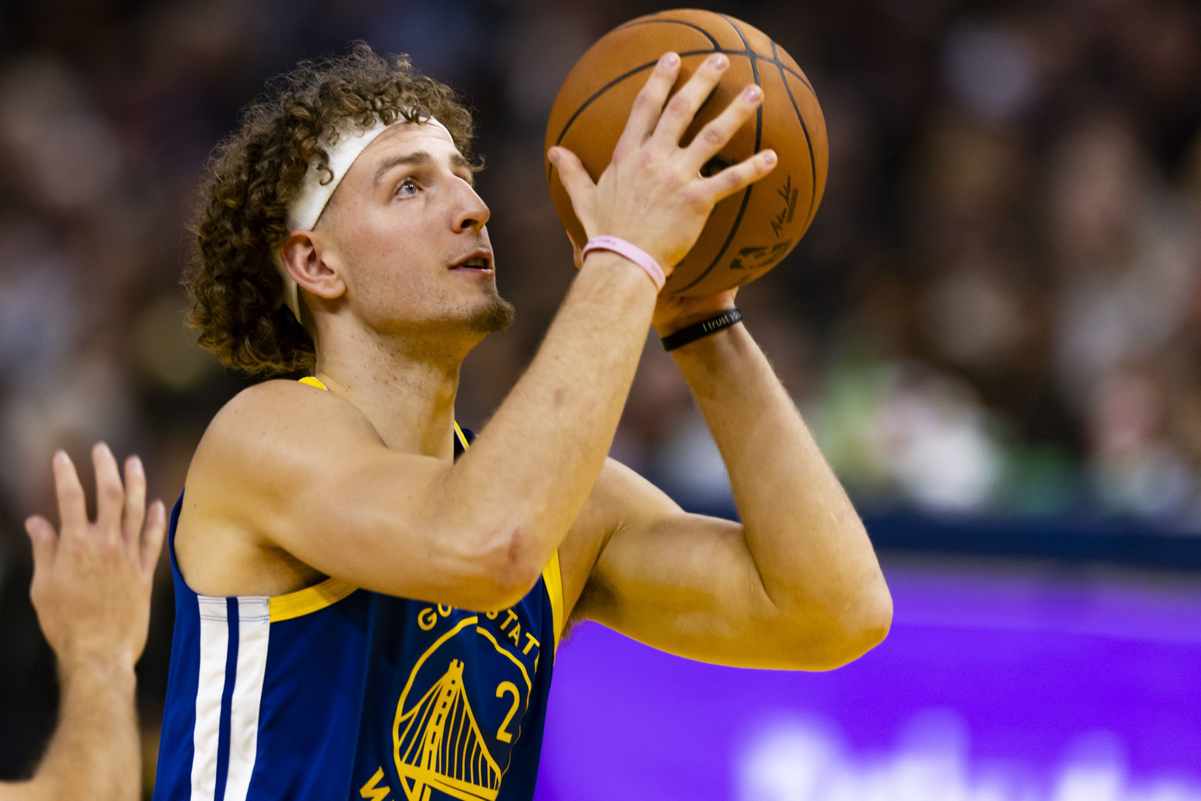 A basketball player wearing a Golden State Warriors jersey prepares to shoot, holding a basketball near his face with focused expression.