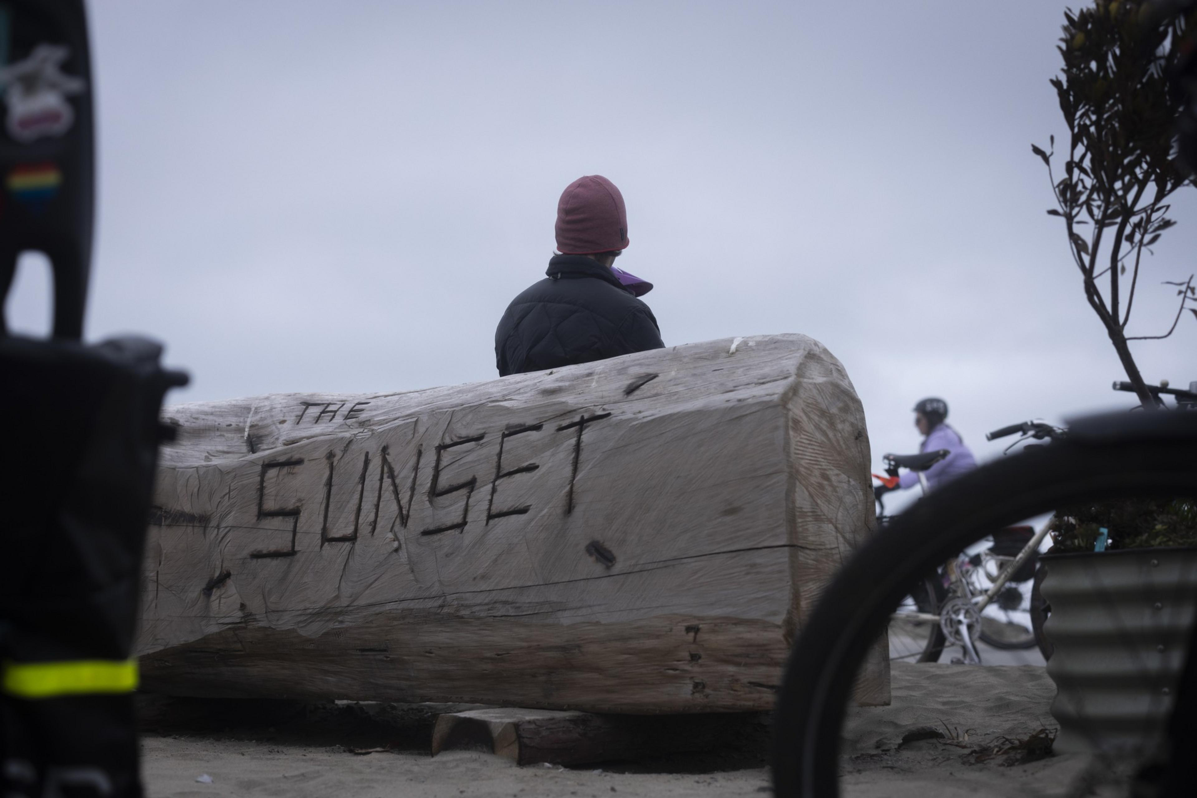A person in a beanie sits on a large log with &quot;THE SUNSET&quot; carved into it, while a cyclist passes by in a cloudy setting.