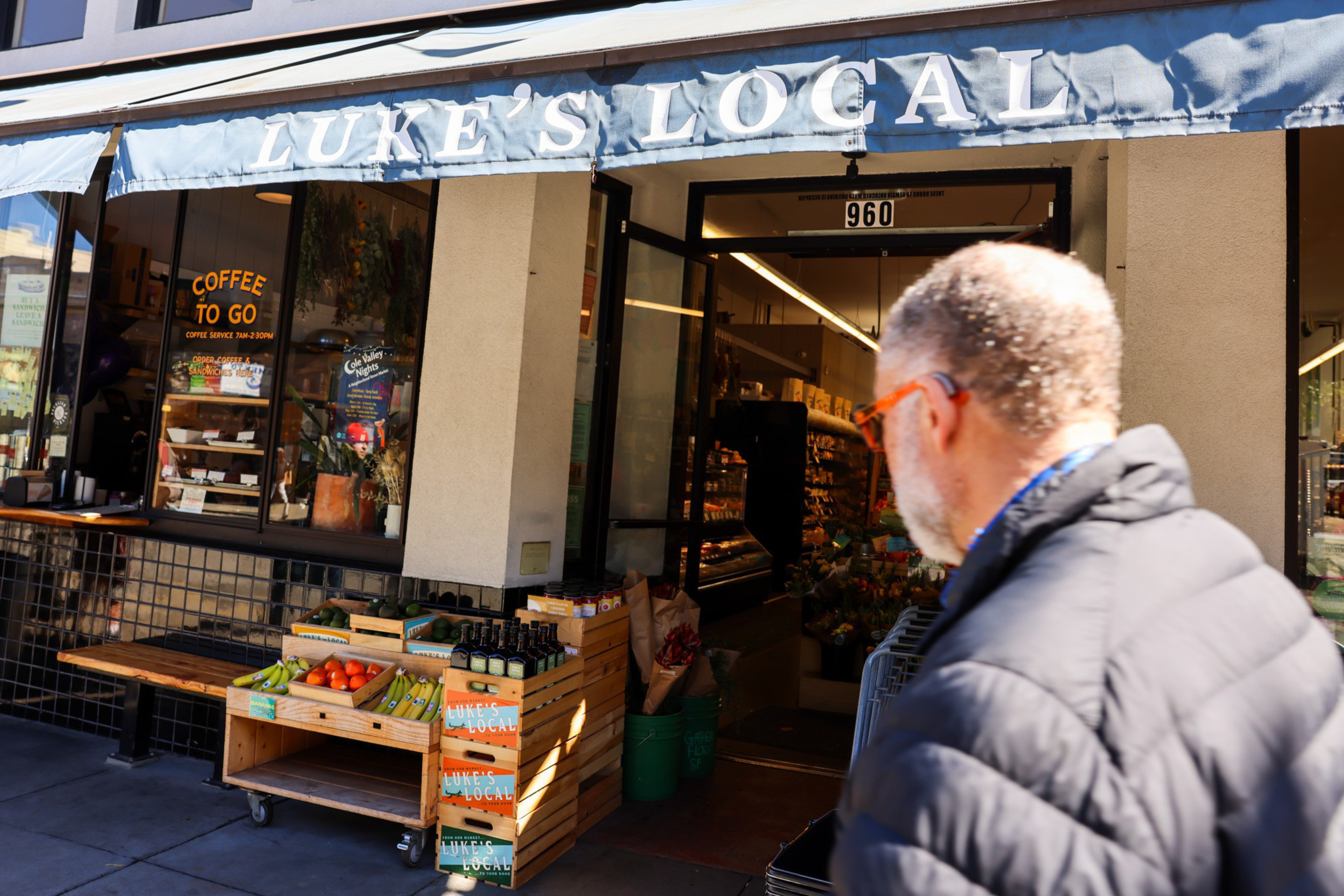 A man with sunglasses walks past a store called &quot;Luke's Local,&quot; displaying fruits, vegetables, and bottled items outside under a blue awning.