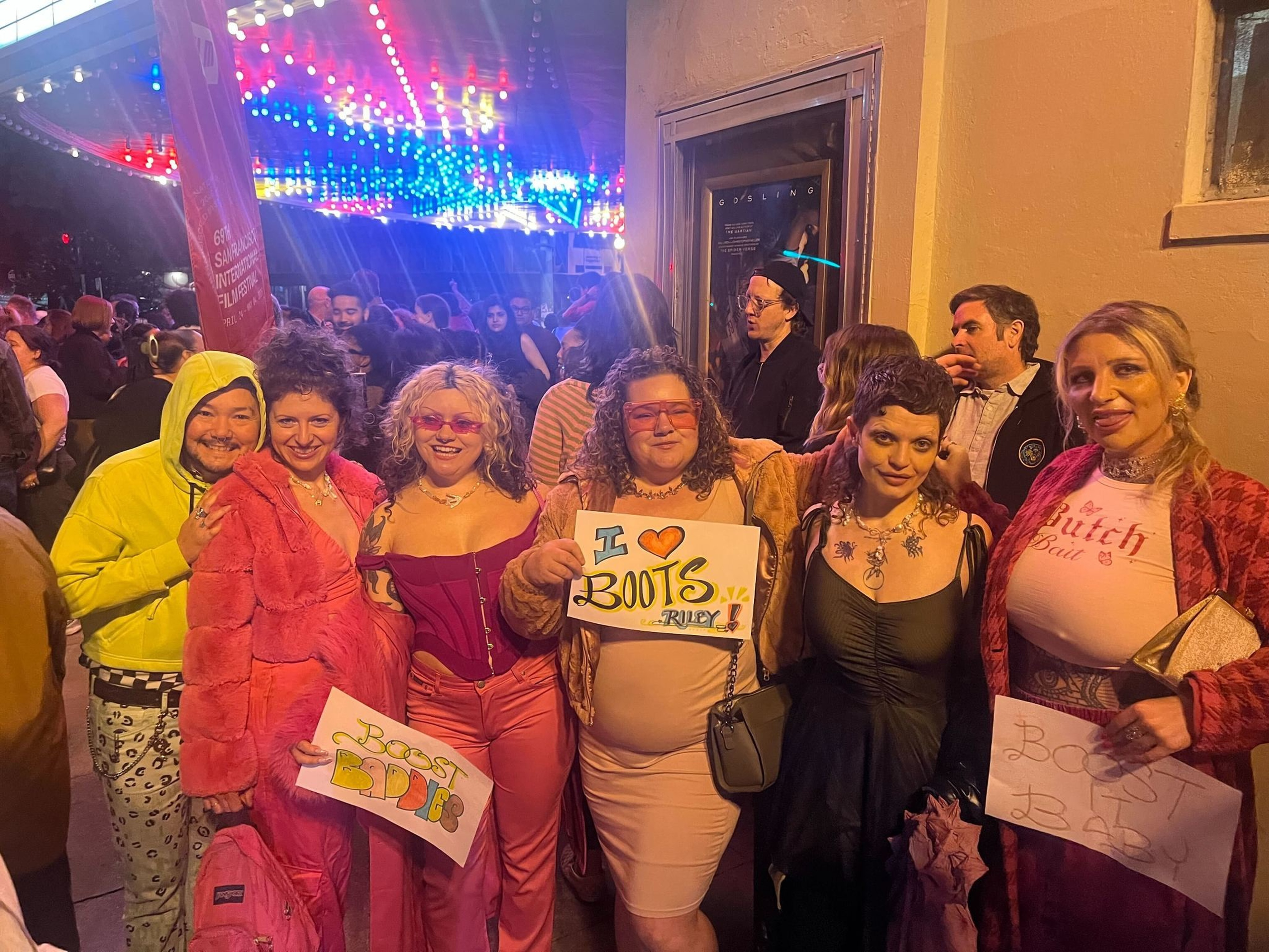 A group of six people dressed in colorful outfits hold signs that say “Boots Babes” and “I ❤️ Boots, Riley” under vibrant, multicolored lights.