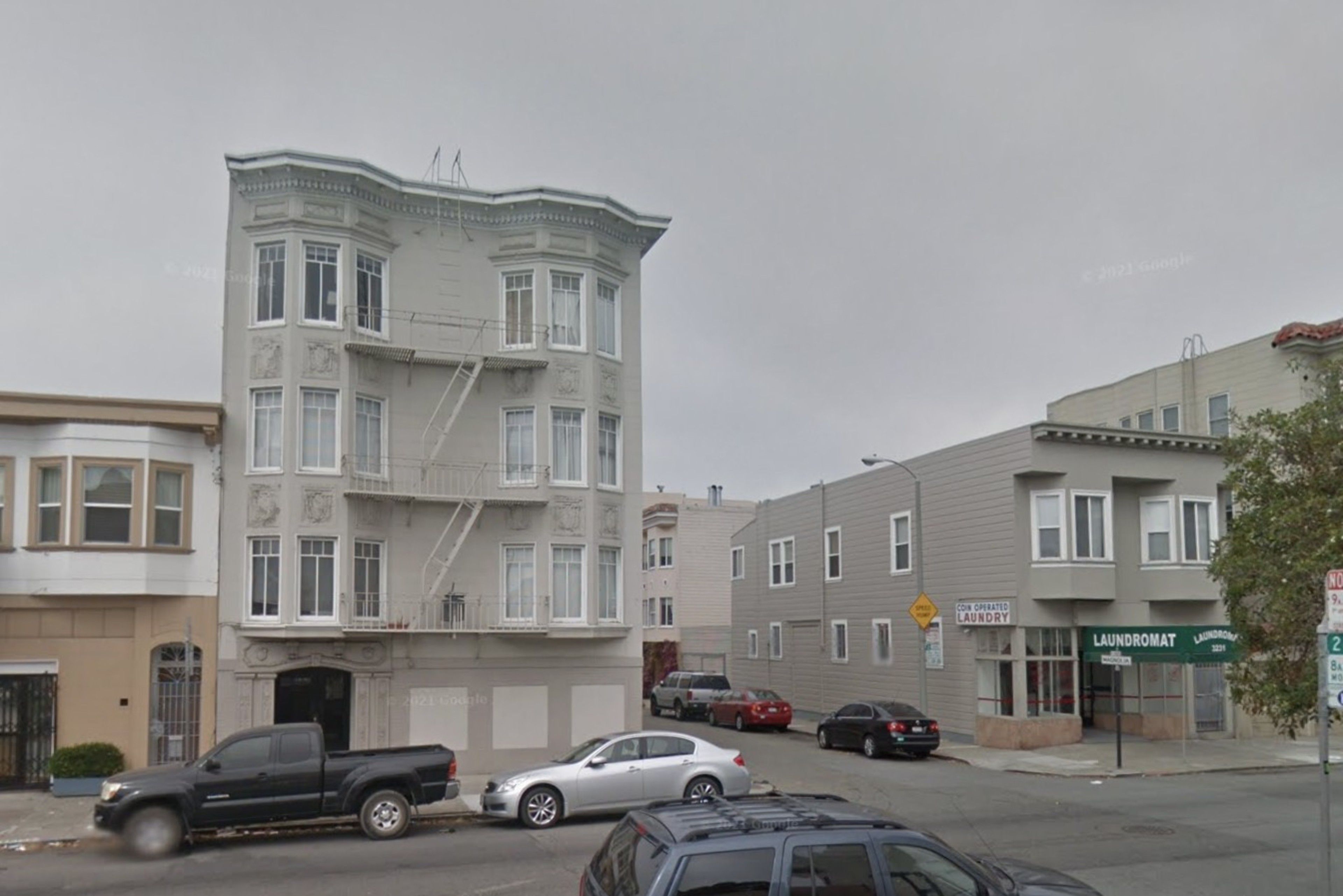 A three-story ornate building with bay windows and external fire escape, next to a single-story laundromat, on a cloudy day.