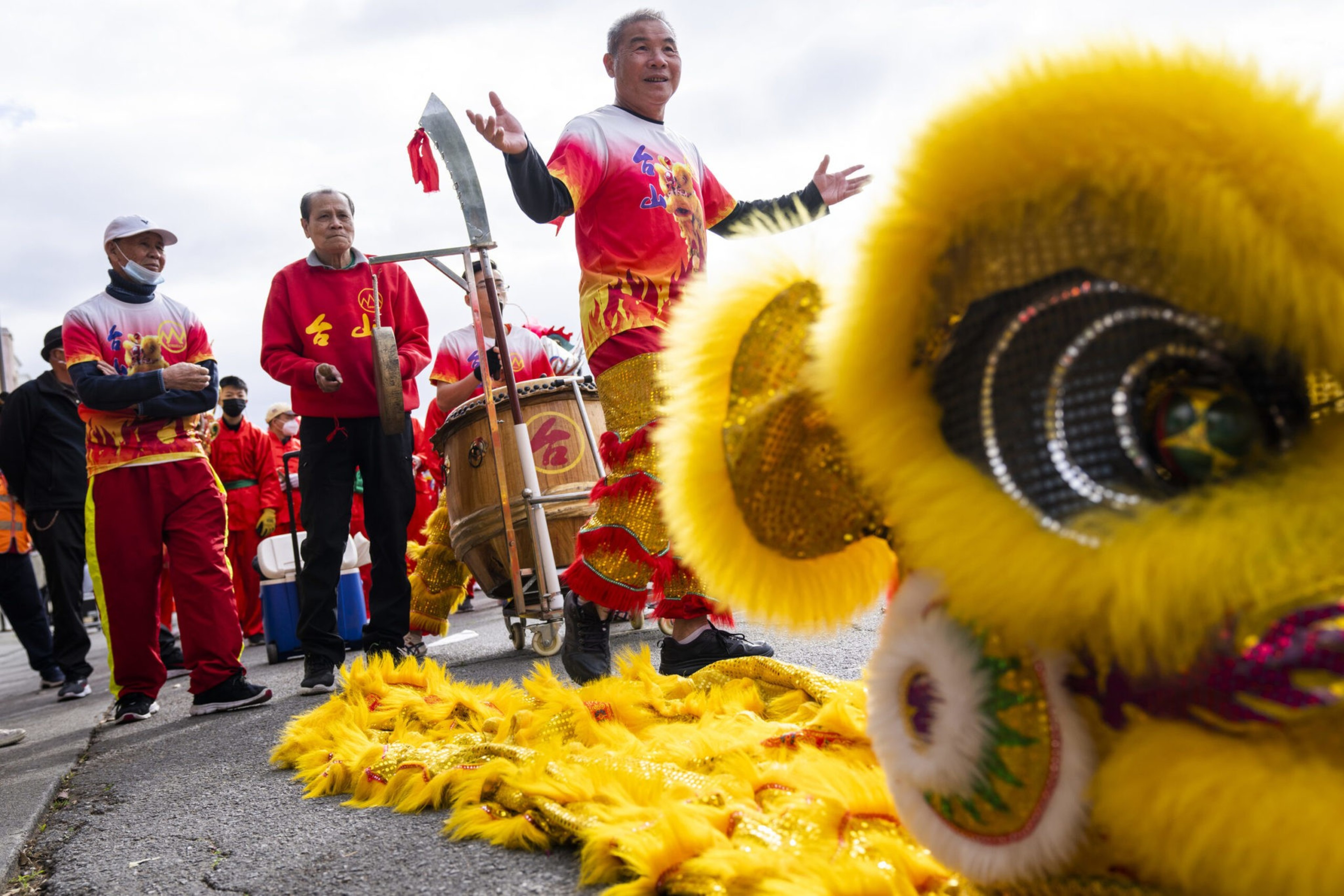 A group performs a traditional lion dance with bright yellow costumes and drums, led by a man in a red and yellow outfit gesturing with open arms.