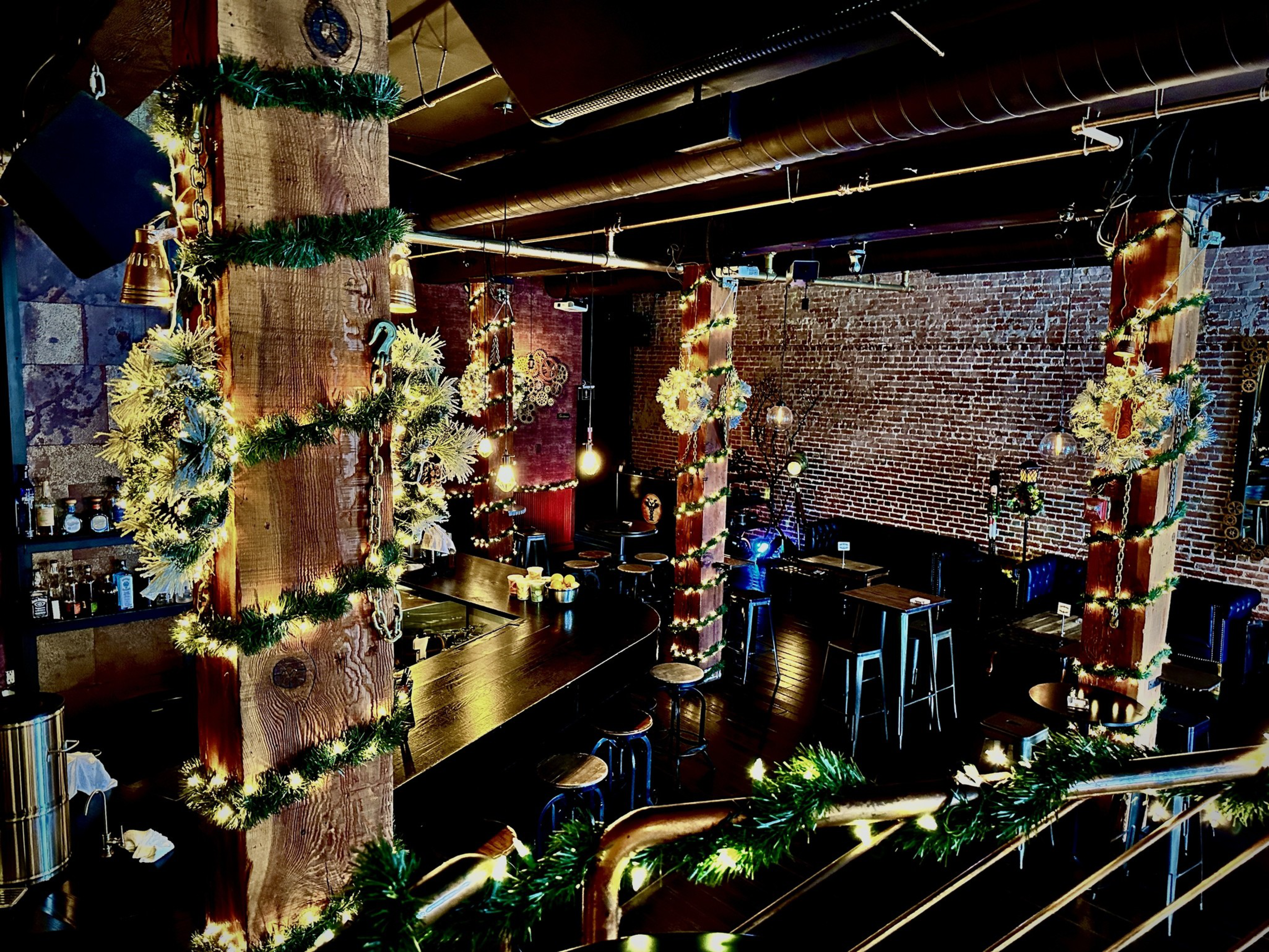 Garlands and lights are wrapped around wooden pillars in an industrial-style bar with exposed brick walls. 