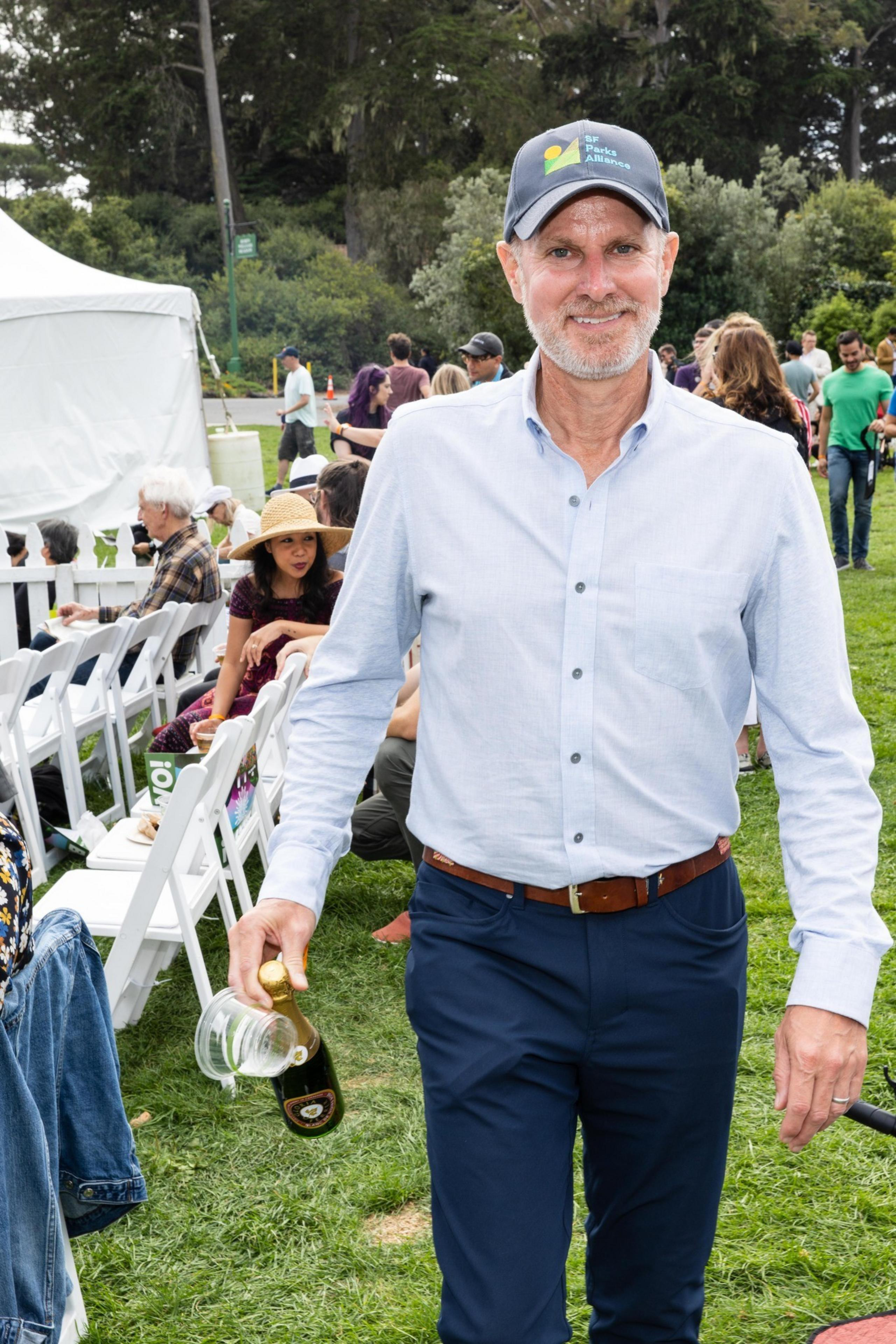 A man in a blue shirt and cap holds a bottle and cups at an outdoor event with people seated and standing on grass, surrounded by trees and a tent.