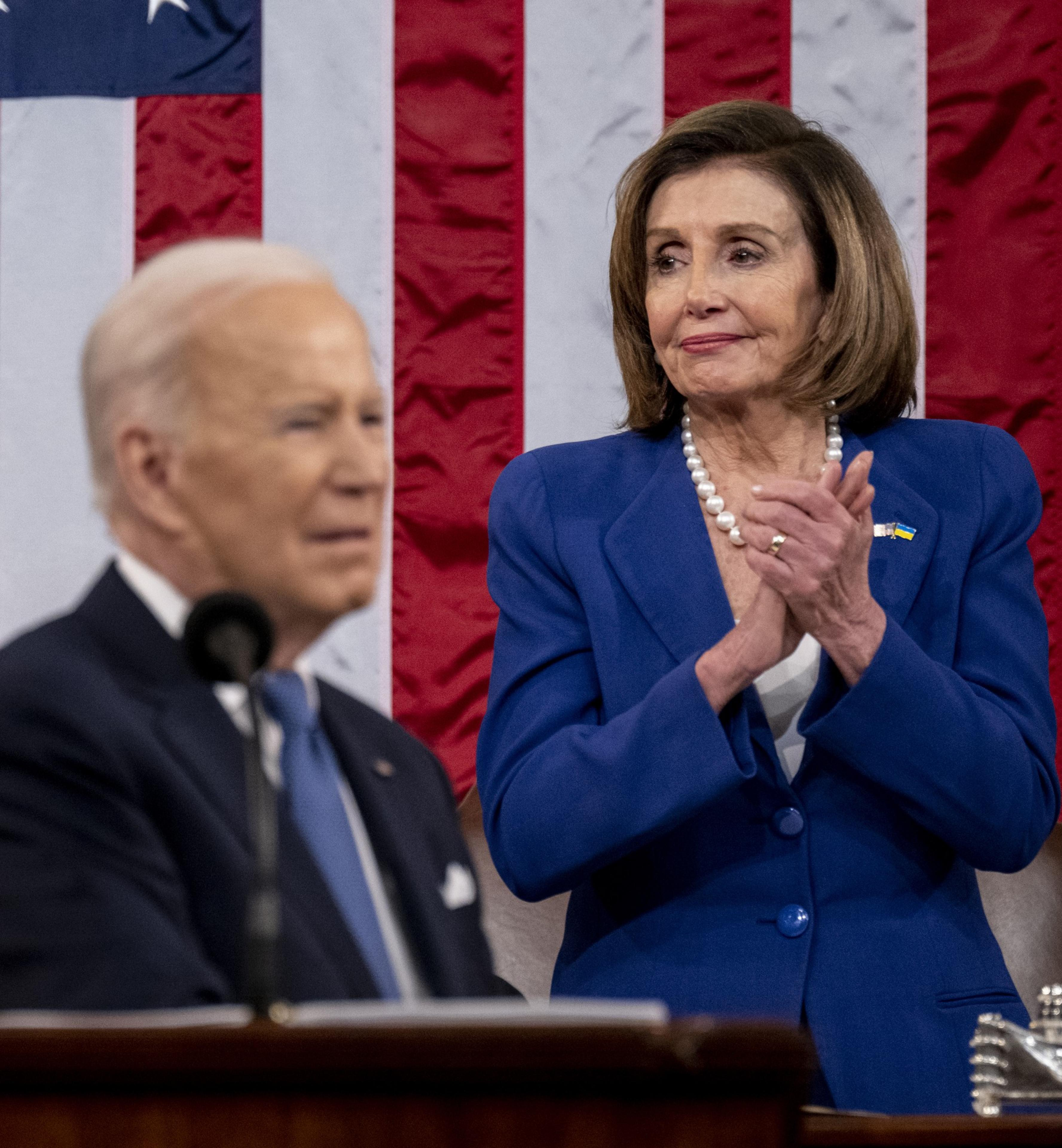 A man speaks from a podium with a microphone, while a woman in a blue blazer and pearl necklace stands behind him, clapping. An American flag is in the background.