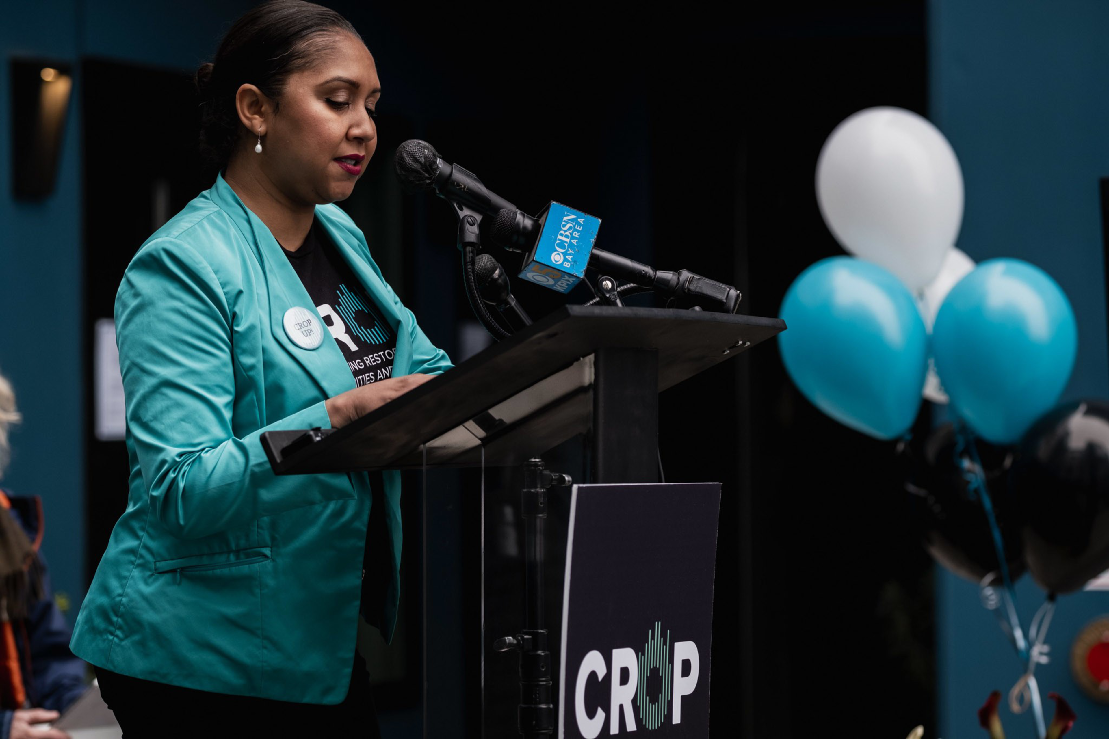A woman in a teal blazer speaks at a podium with a "CROP" sign, with blue and white balloons in the background and a CBSN microphone attached.