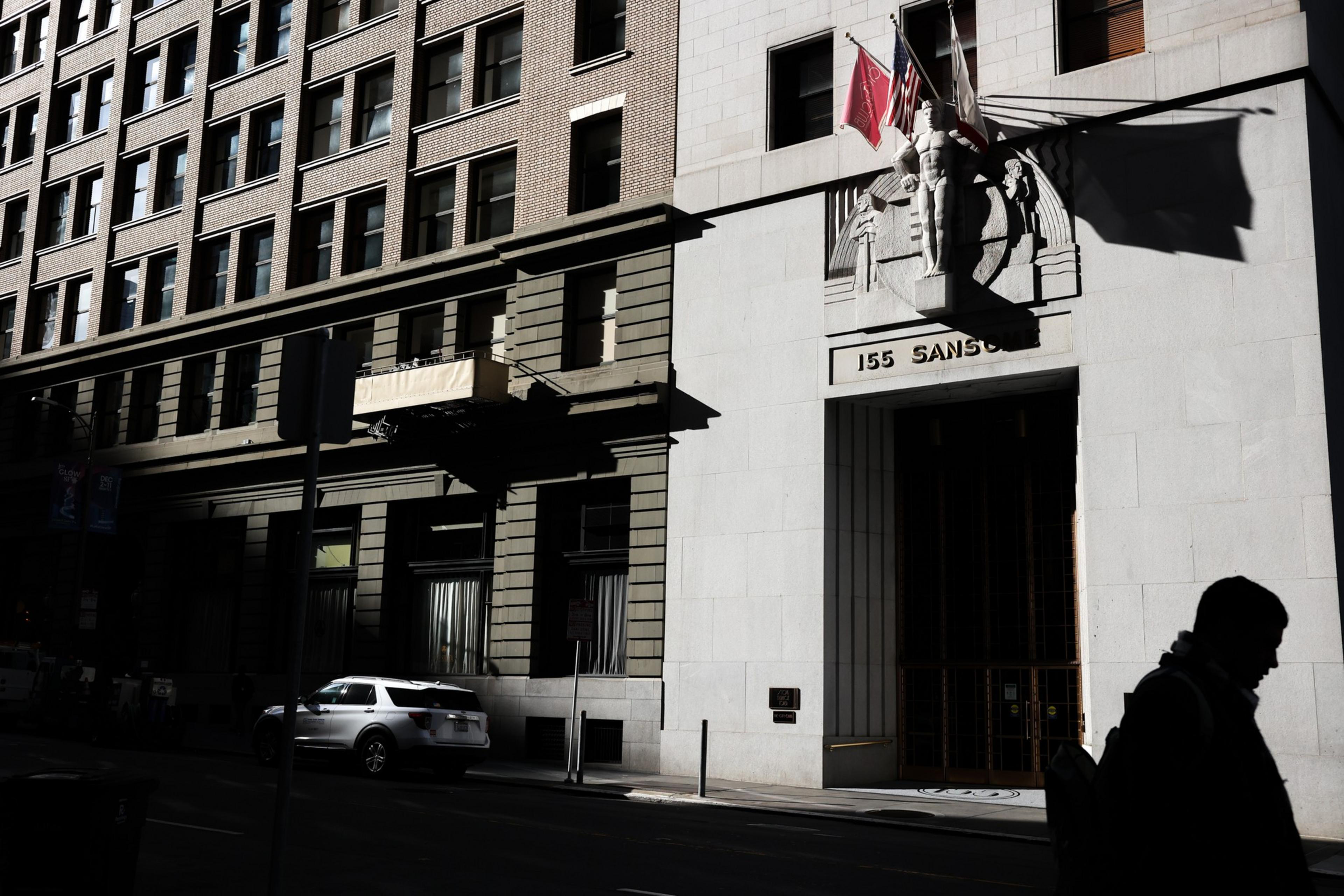 A building entrance marked "155 Sansom" with flags above and a shadowed person walking past in front of the sunlight-cast facade.