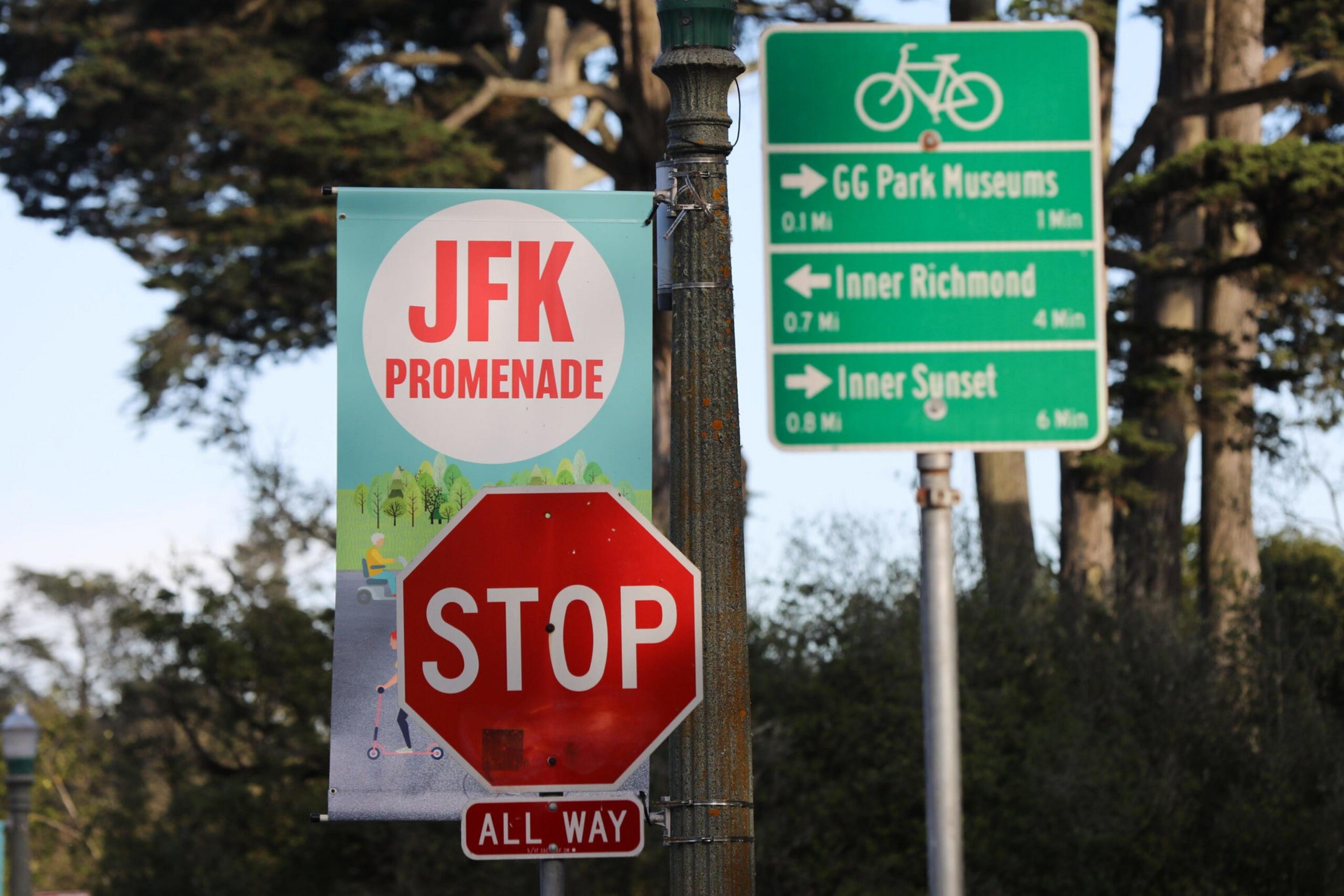 A red stop sign with an "ALL WAY" plaque below stands in front of a "JFK Promenade" banner and a green bike route sign showing directions to GG Park Museums, Inner Richmond, and Inner Sunset.