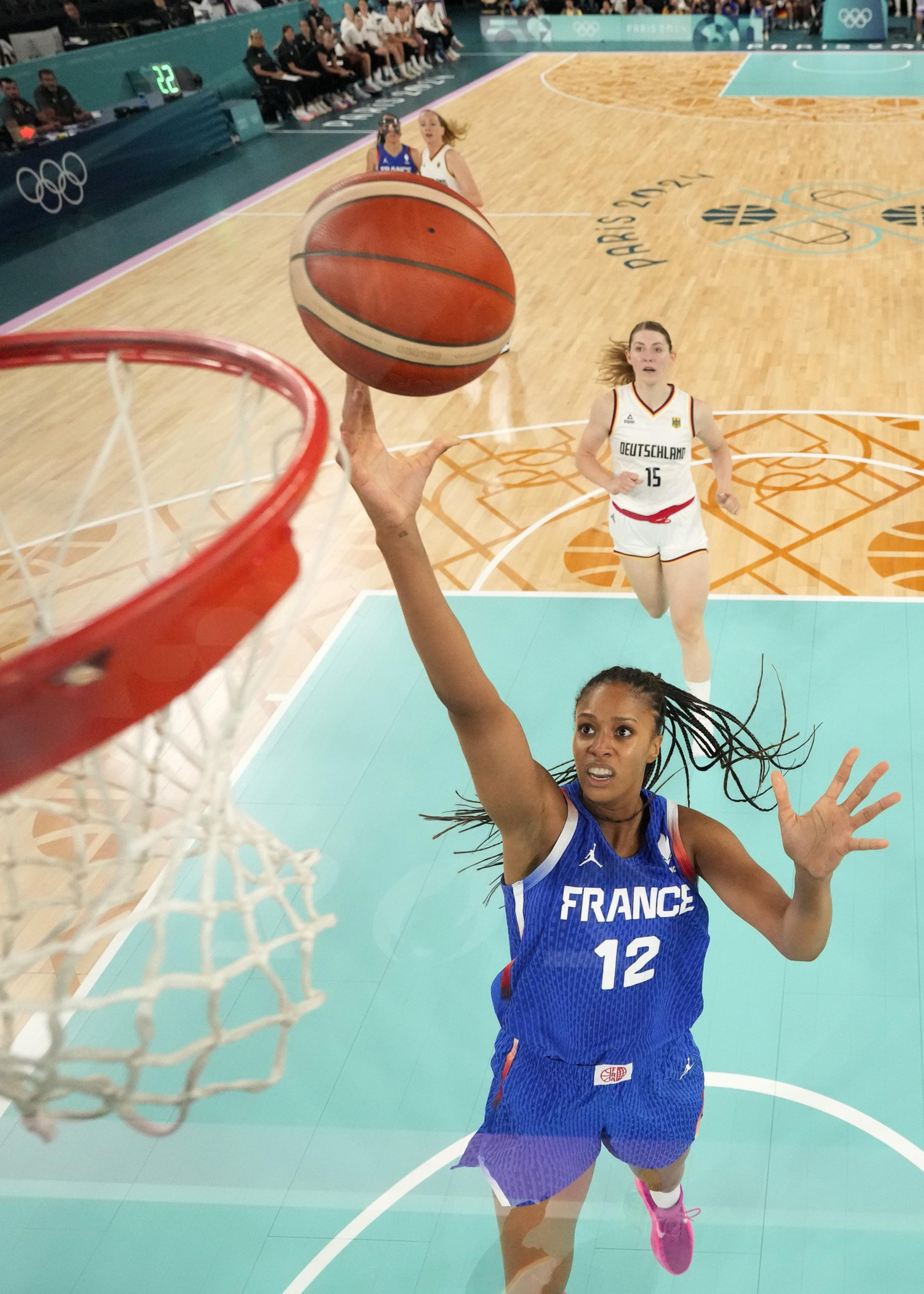 A basketball player in a France jersey is leaping to score, with a Germany player behind her on a brightly colored court, viewed from above the hoop.