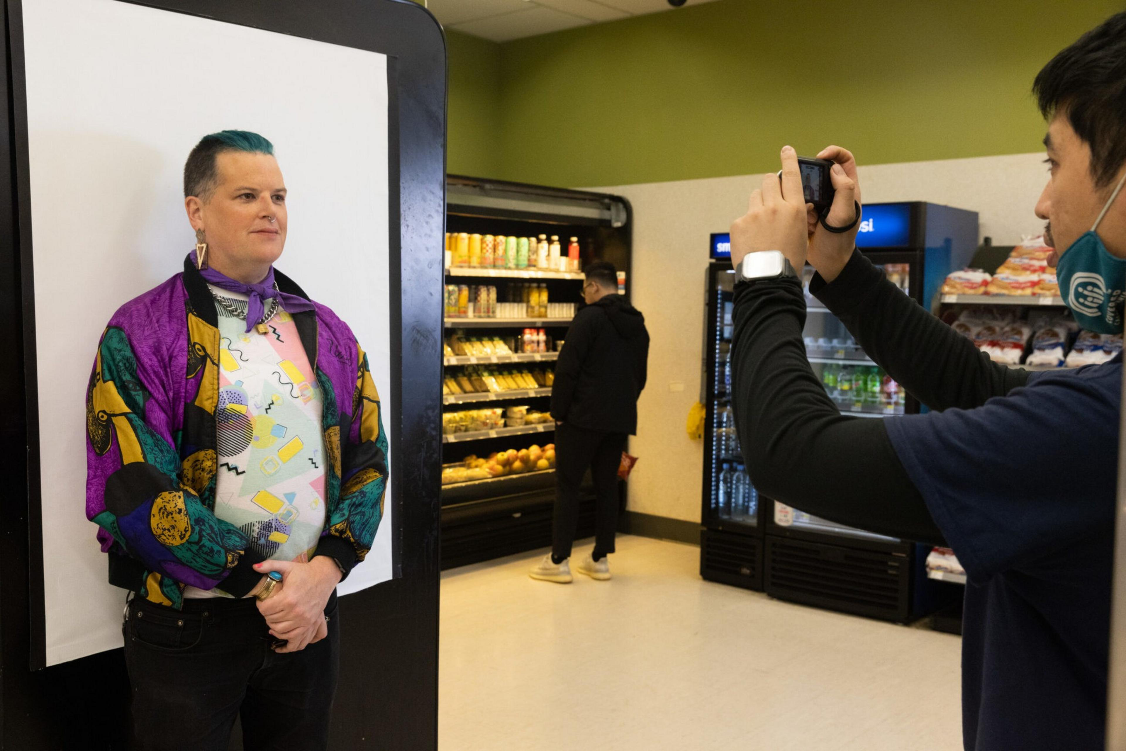 A person with short teal hair and a colorful jacket poses for a photo in a store while another person takes the picture, and a third person shops nearby.