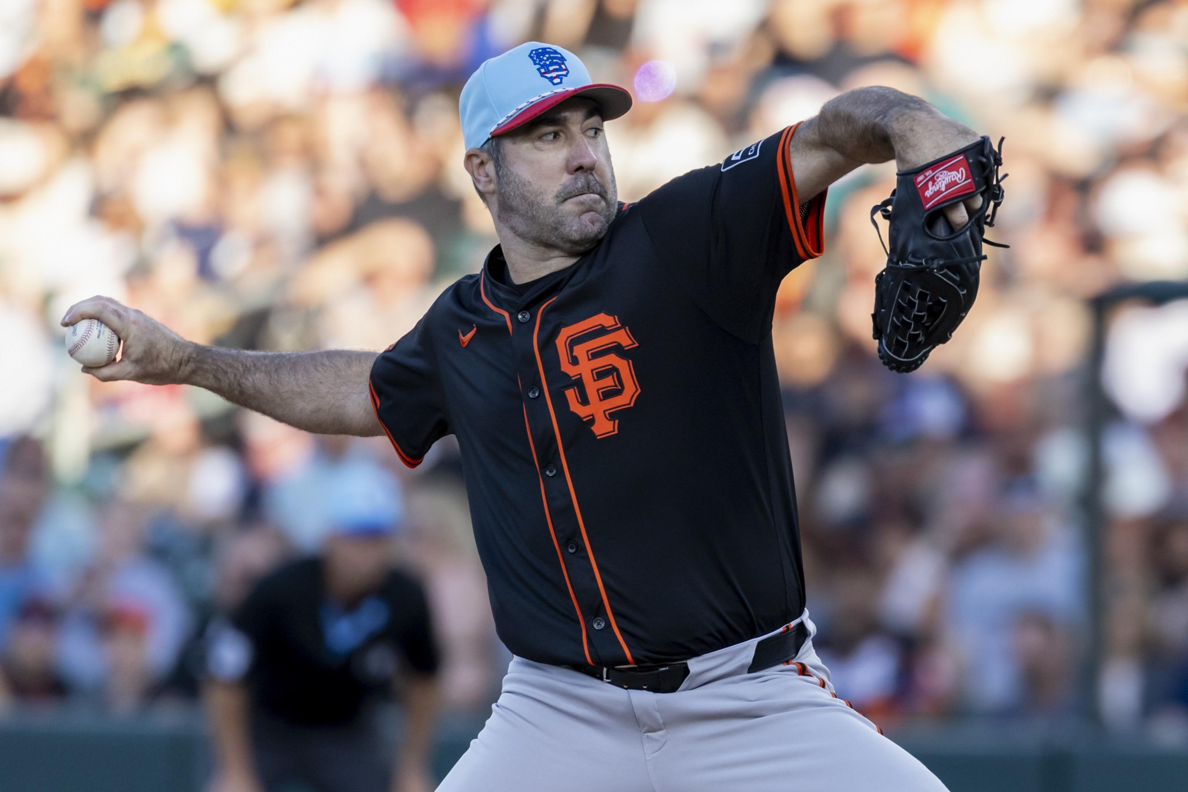 A baseball player in a black jersey with an orange "SF" emblem is mid-pitch on a field. He wears a light blue cap, glove, and holds a baseball. The crowd is blurred behind him.
