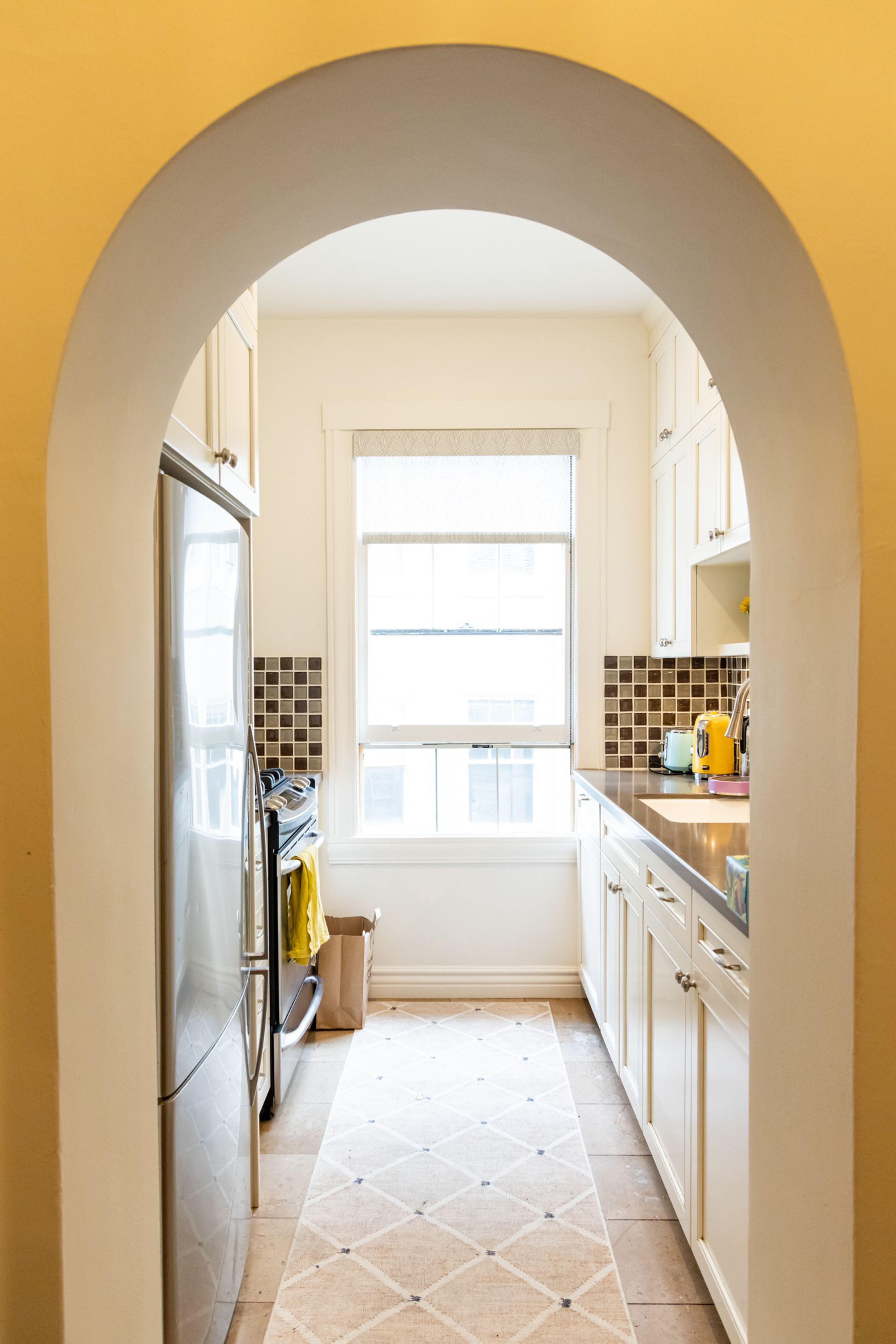 A narrow kitchen with white cabinets, stainless steel appliances, a window at the end, a beige rug on the floor, and a brown mosaic tile backsplash.